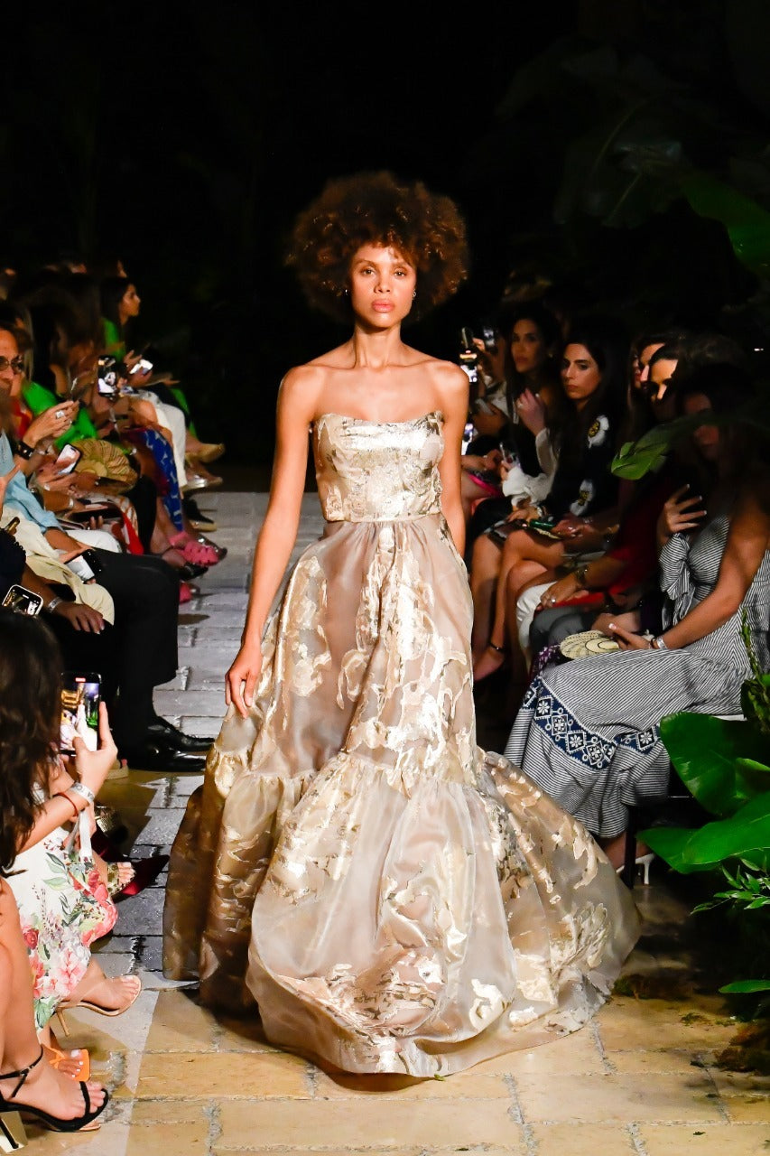 Model walking down a runway in a strapless, gold-patterned gown, surrounded by seated audience.