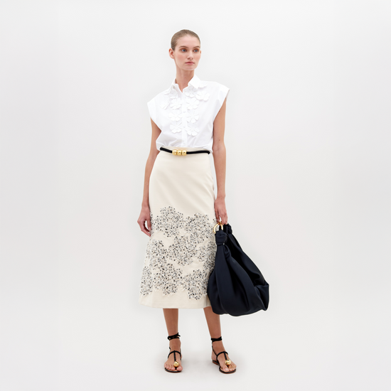 A woman stands wearing a white patterned jacket, Atira Skirt Beige Black Embroidered Flowers, gold belt, and gold-embellished sandals. She holds a beige handbag against a plain background. Don't miss out—pre-order now for the November 15th, 2024 ship-by date!