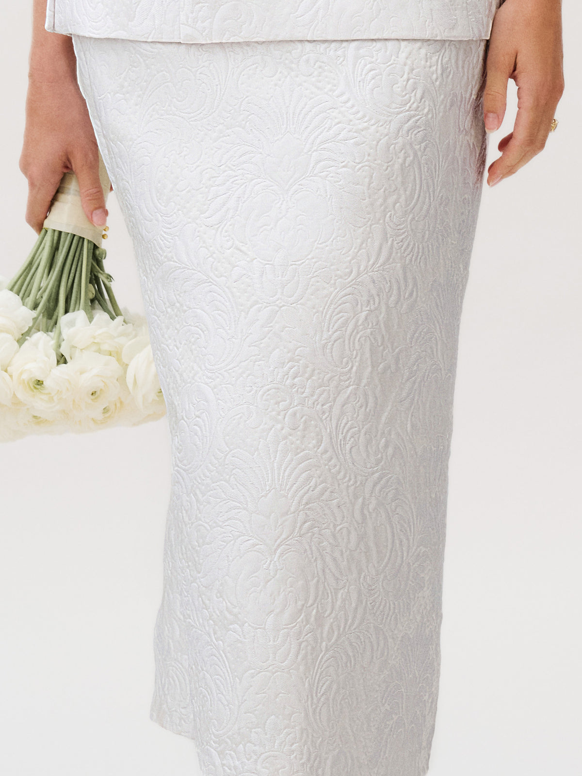 A person stands against a plain background wearing the Atira Skirt, an elegant, textured white piece crafted from luxurious Italian fabric, while holding a bouquet of white flowers in their left hand.