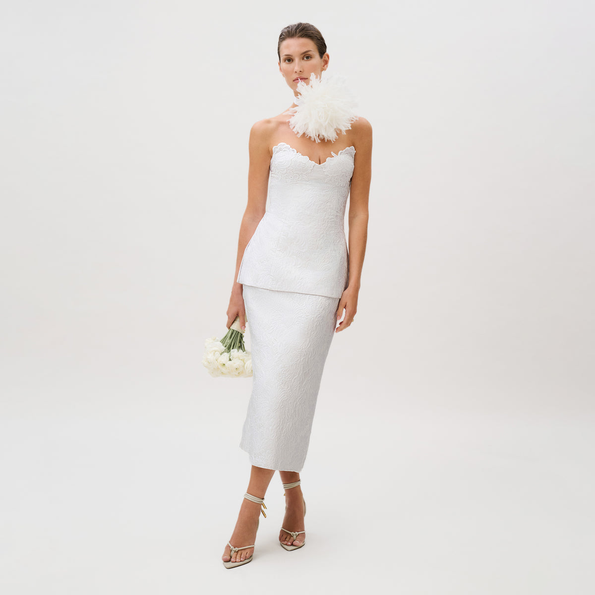 A woman stands against a plain background, holding white flowers and wearing the Atira Skirt—crafted from luxurious Italian fabric in a strapless, white design with a dramatic feather detail on one shoulder.