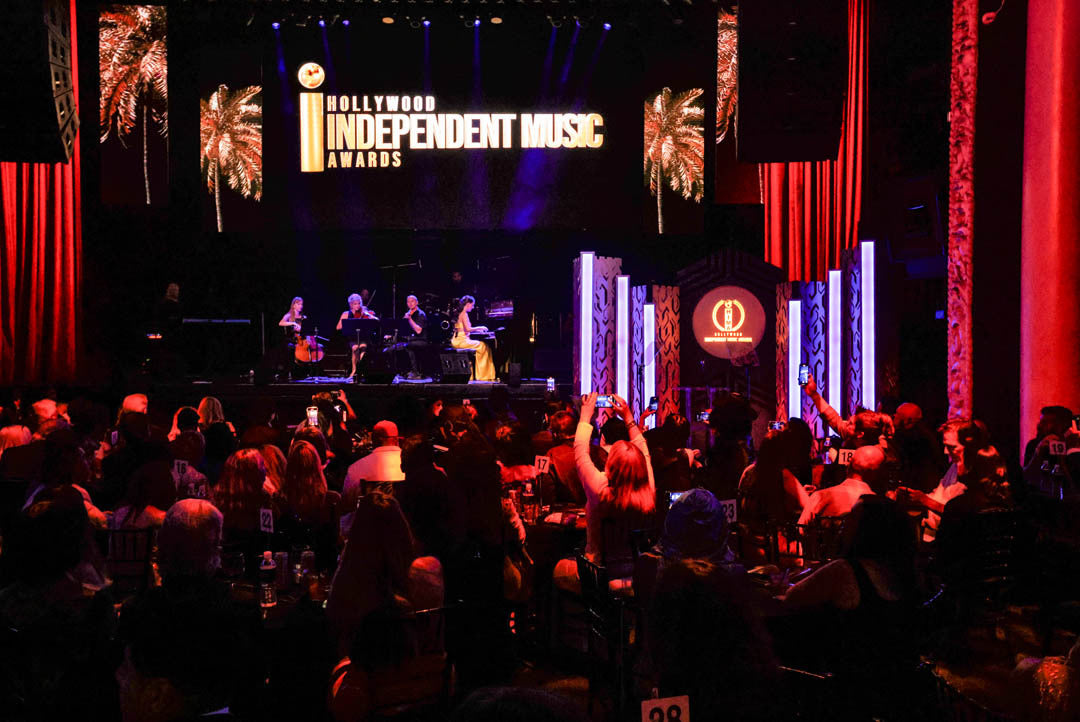 Audience watches a piano performance on stage at the Hollywood Independent Music Awards, with "Hollywood Independent Music Awards" sign and palm trees in the background.