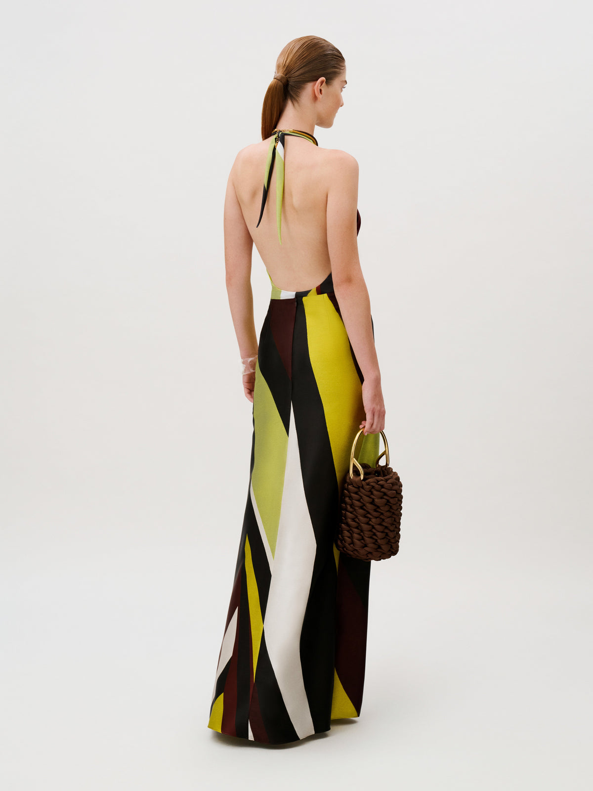 Back view of a person wearing an Aura Skirt floor-length silk halter-neck dress with a geometric pattern in lime, black, and white, holding a brown woven handbag.