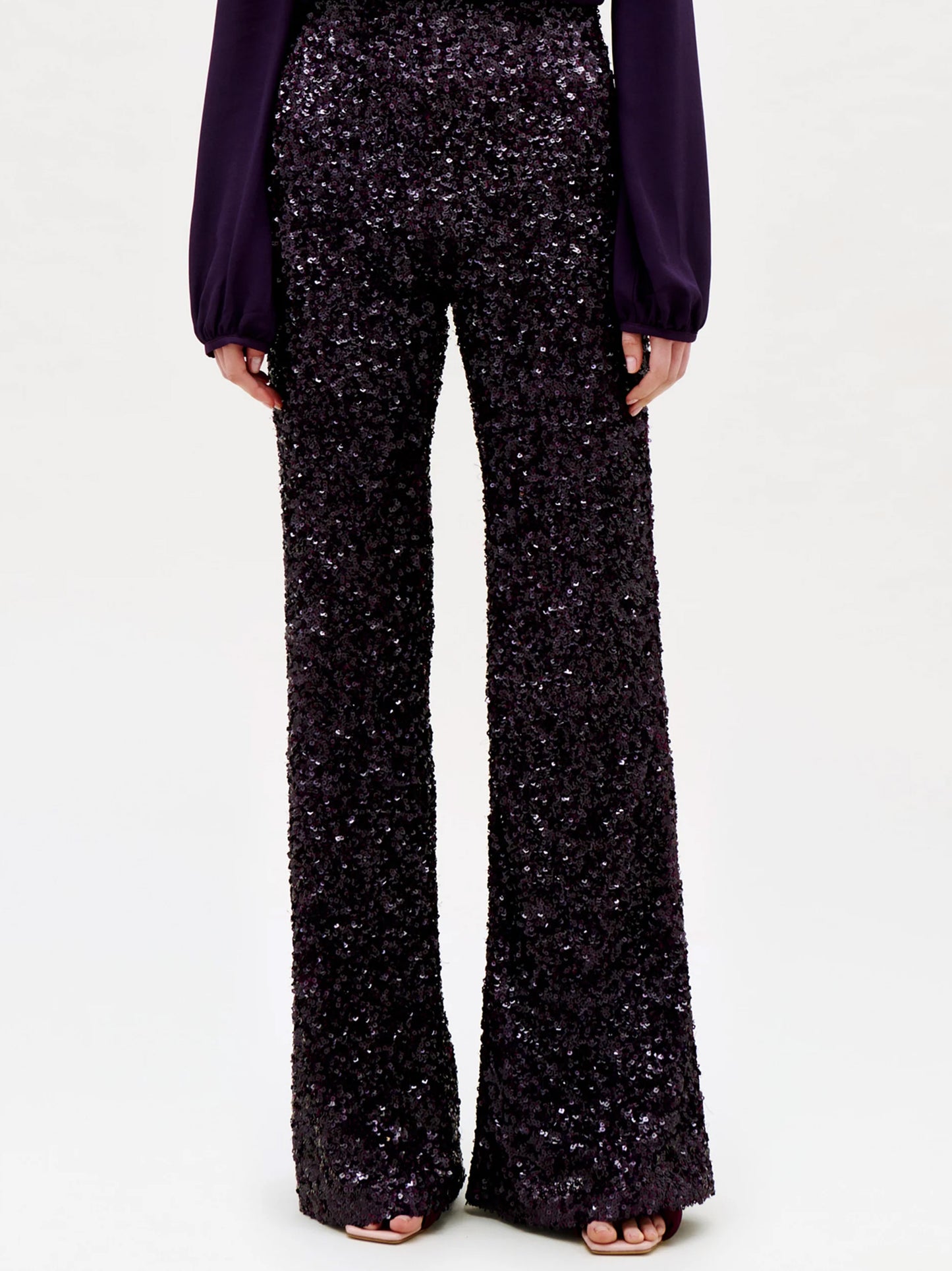 A person wearing the Avellino Pant—a pair of high-waisted, wide-leg pants crafted in black sequins—paired with a long-sleeve dark purple blouse, standing against a plain light background.