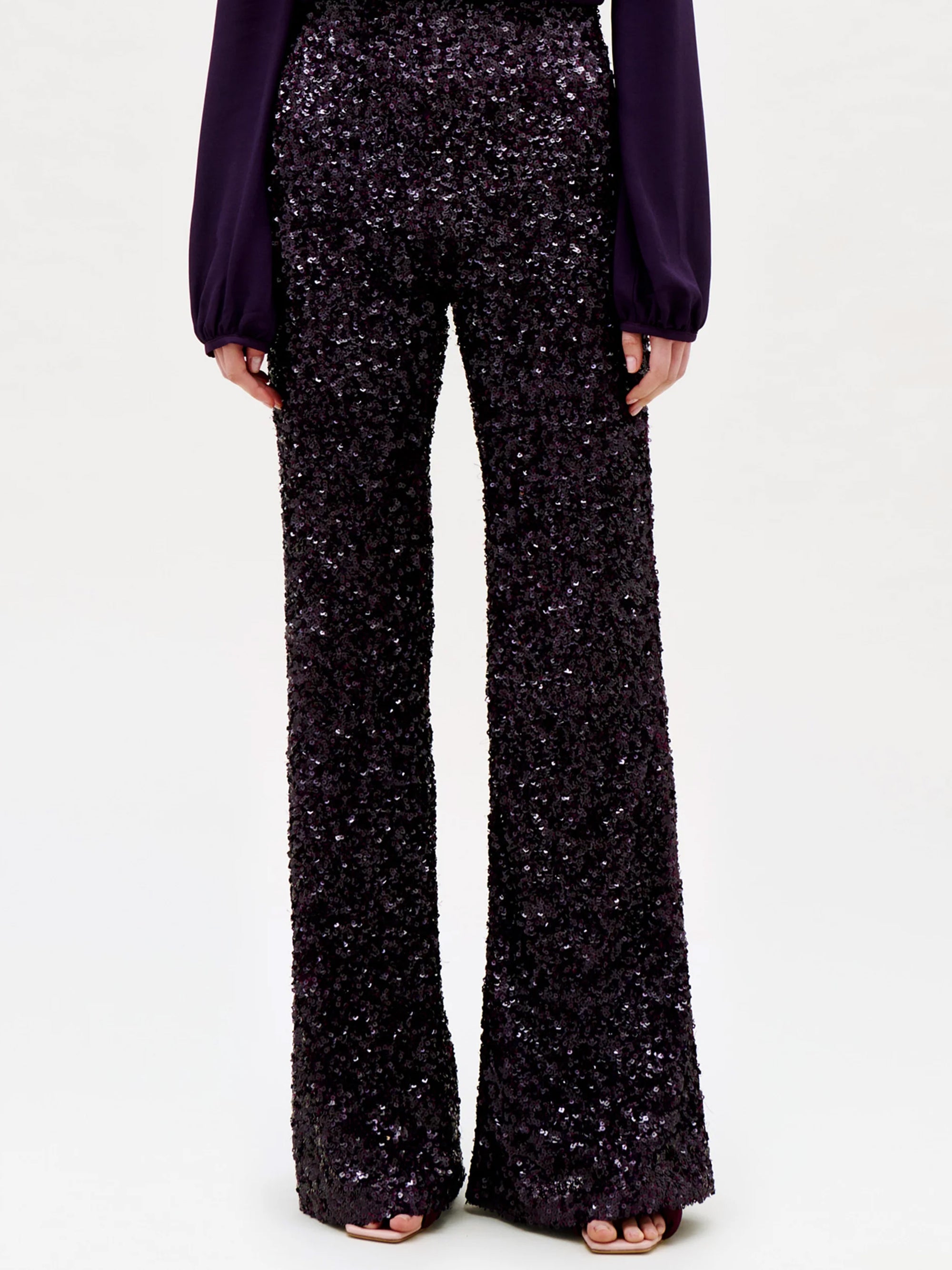 A person wearing the Avellino Pant—a pair of high-waisted, wide-leg pants crafted in black sequins—paired with a long-sleeve dark purple blouse, standing against a plain light background.