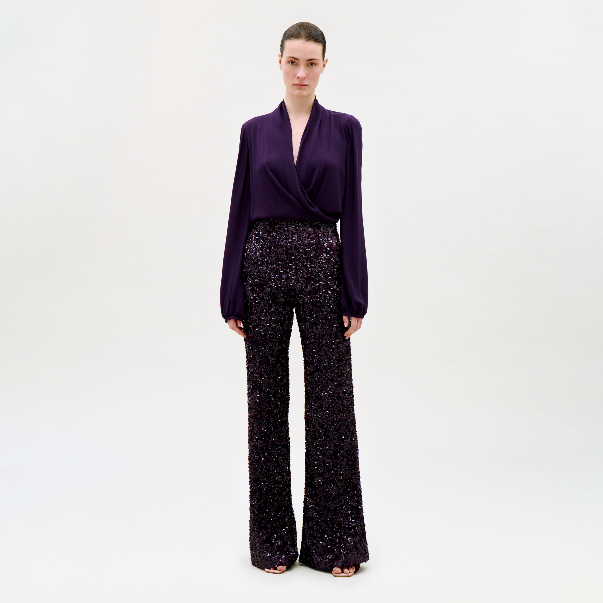 A woman with her hair tied back and a neutral expression stands against a light background, wearing the Avellino Pant—high-waisted black sequin wide-leg pants—with a deep purple wrap blouse and black open-toe shoes.