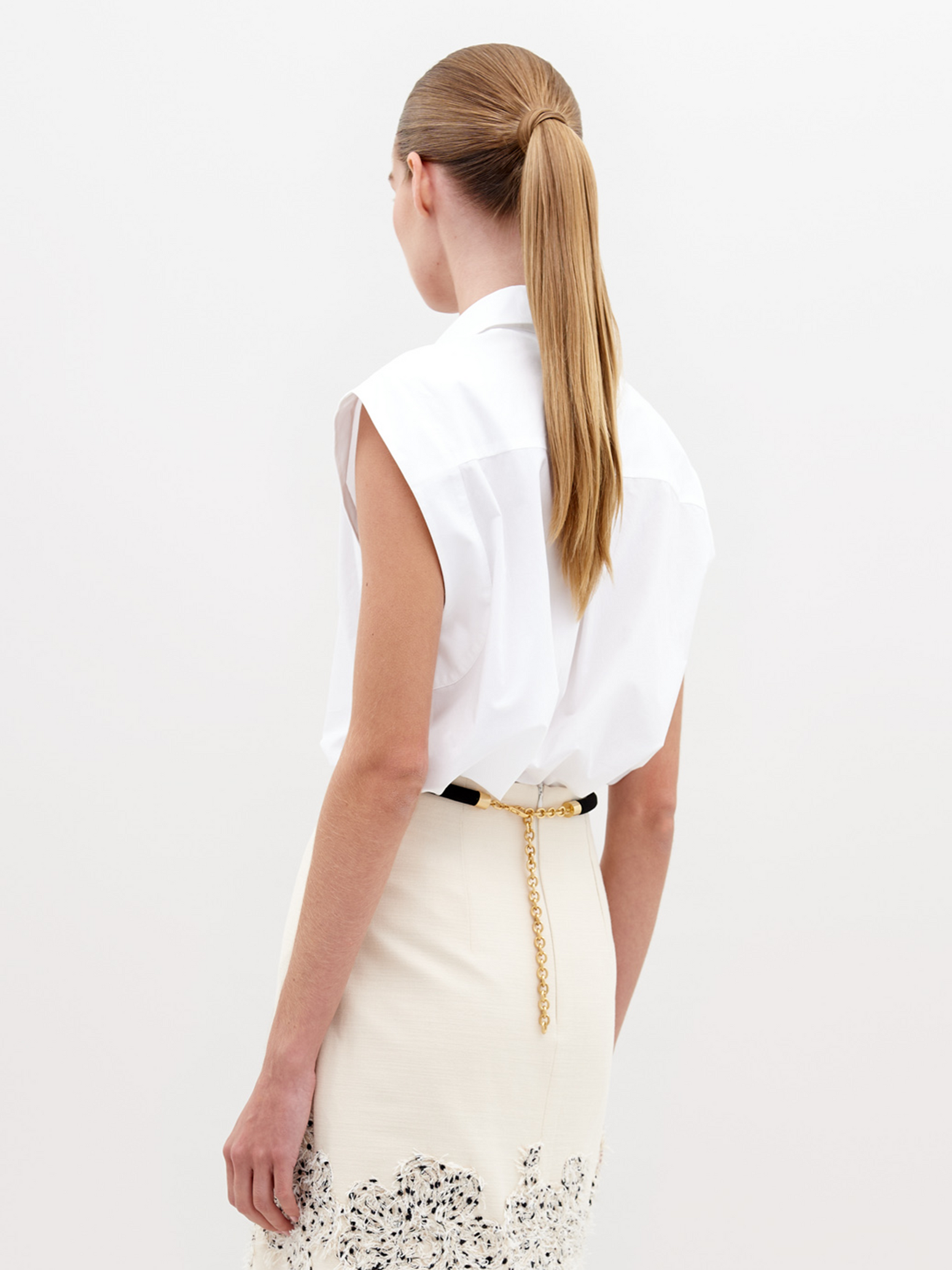 The individual is dressed in an Azalea Blouse, a white sleeveless top with floral appliqué, paired with a cream skirt featuring a patterned hem, and holds a black bag against a plain background.