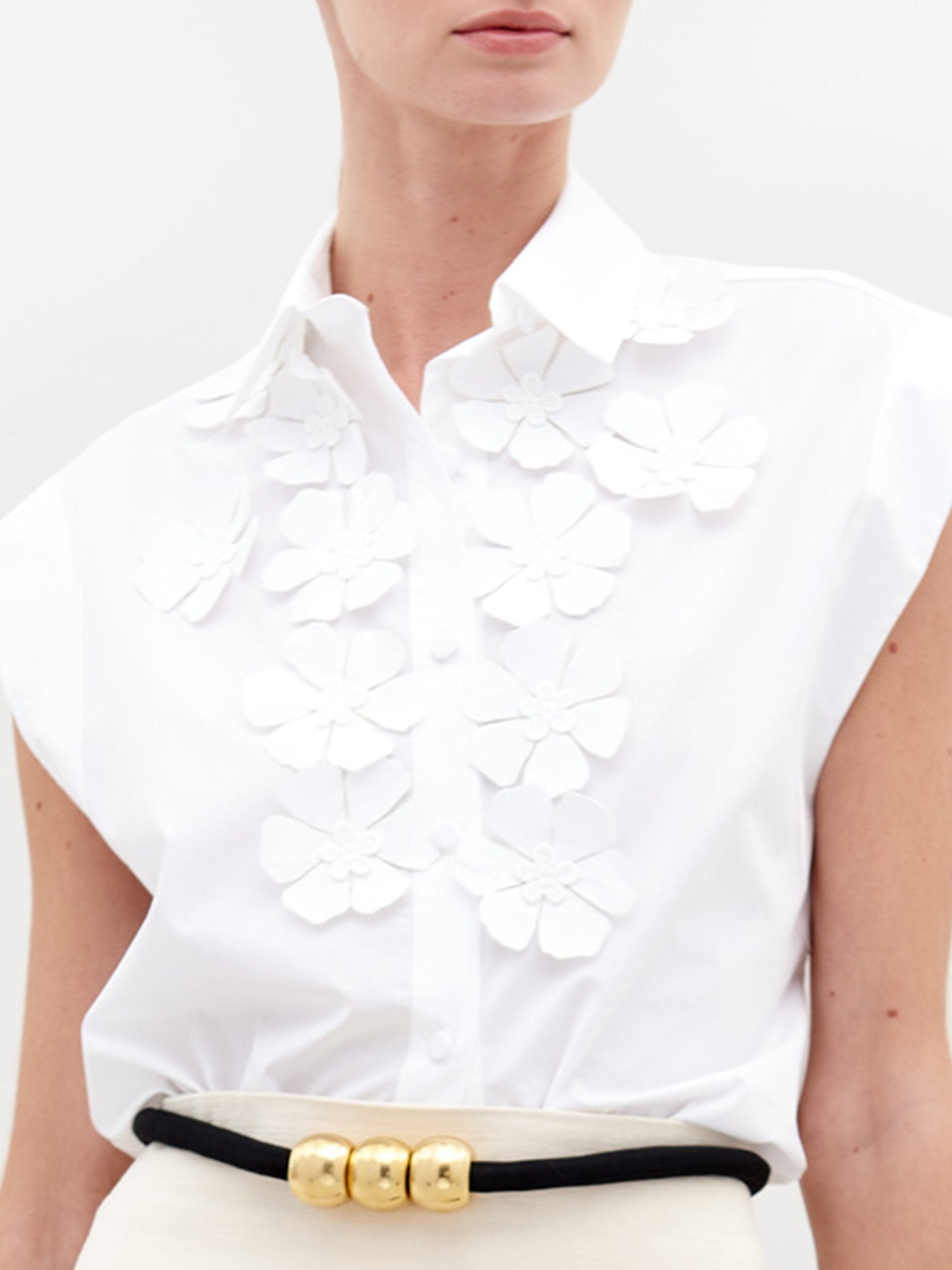 The individual is dressed in an Azalea Blouse, a white sleeveless top with floral appliqué, paired with a cream skirt featuring a patterned hem, and holds a black bag against a plain background.