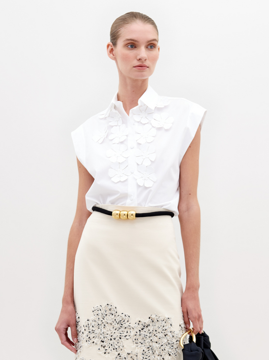 A person wearing the Azalea Blouse in white, which is sleeveless and adorned with appliqué floral detailing, paired with a skirt featuring a black belt and gold accents.