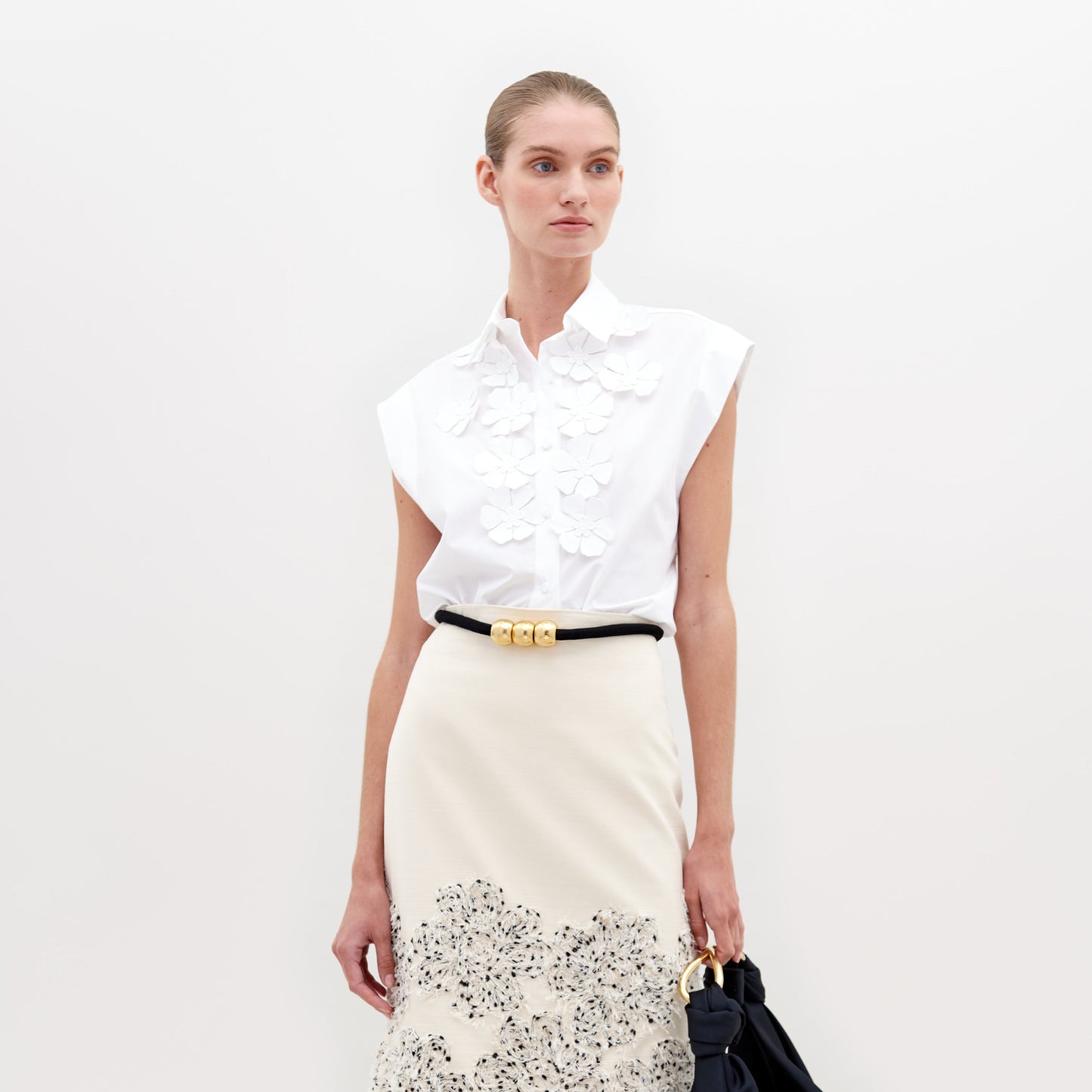 The individual is dressed in an Azalea Blouse, a white sleeveless top with floral appliqué, paired with a cream skirt featuring a patterned hem, and holds a black bag against a plain background.