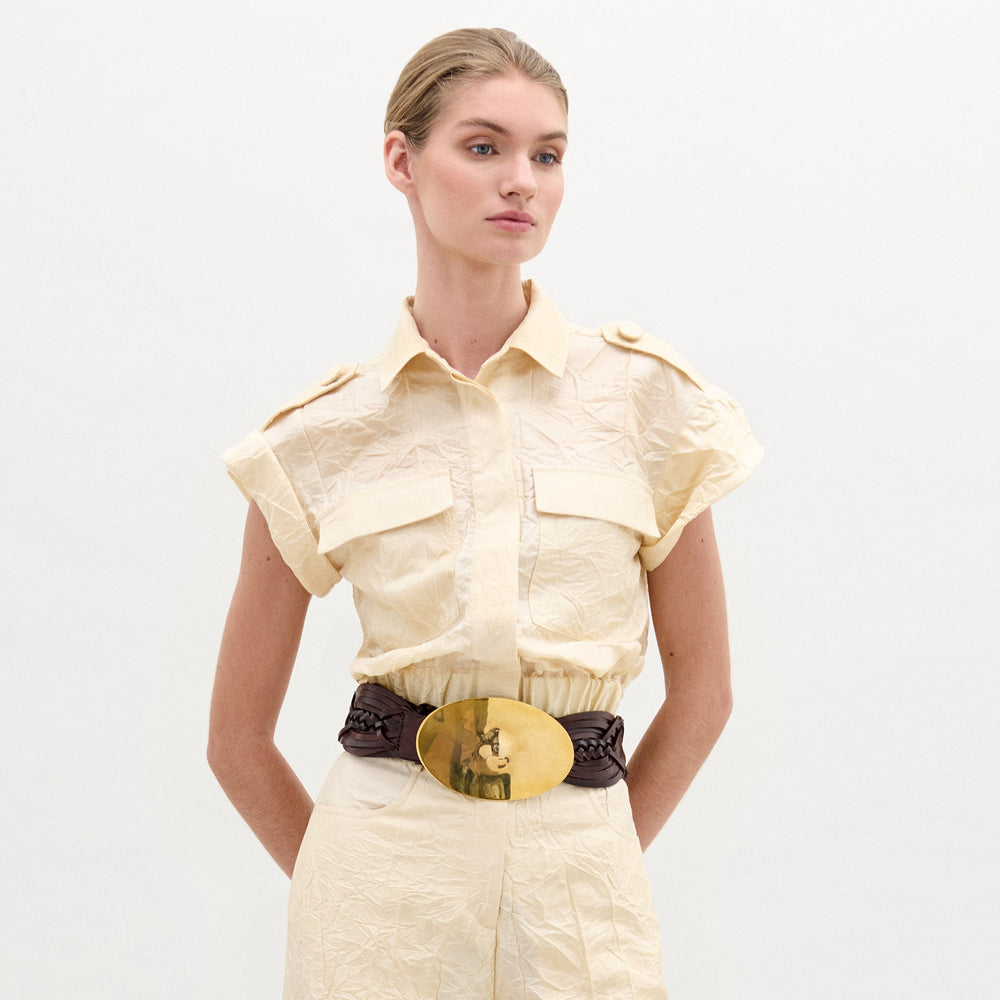 A woman wears the Bara Blouse, a short-sleeved, light beige utility piece made from Italian fabric with front and chest pockets and a wide brown belt with a gold buckle. She stands against a plain white background.