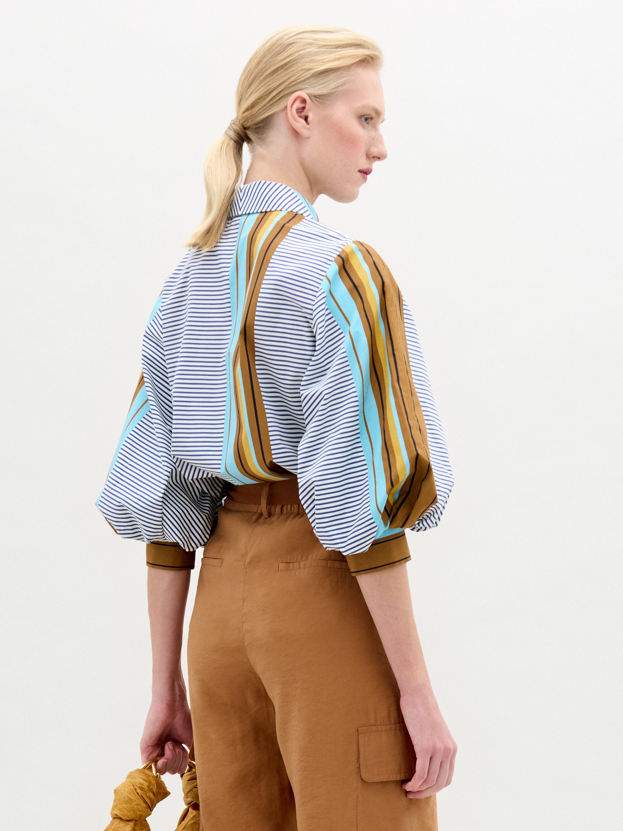 A woman stands against a plain background, wearing the Belkis Blouse with puffed sleeves and oversized tan high-waisted pants, holding a brown bag in her left hand.