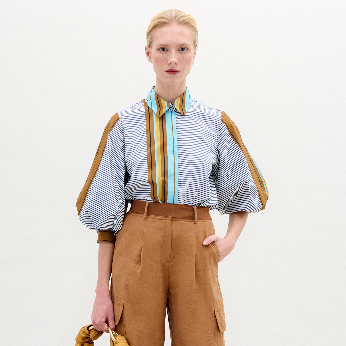 A person stands against a plain background wearing the oversized fit Belkis Blouse with balloon sleeves and stripes, paired with tan high-waisted pants, and holding a tan accessory in their left hand.