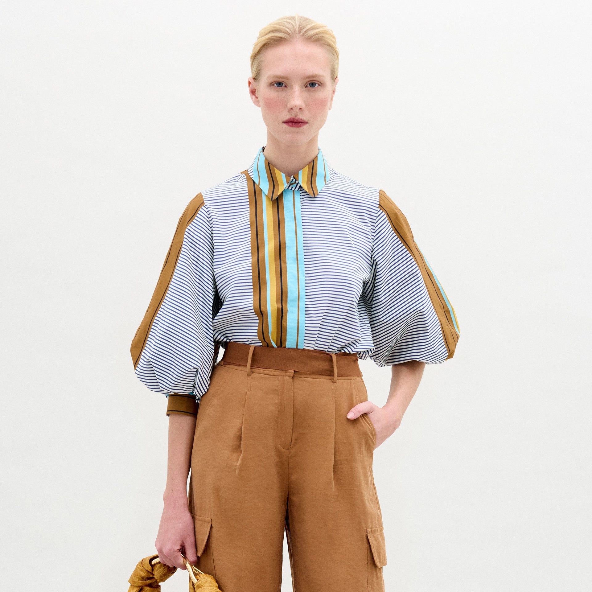 A person stands against a plain background wearing the oversized fit Belkis Blouse with balloon sleeves and stripes, paired with tan high-waisted pants, and holding a tan accessory in their left hand.