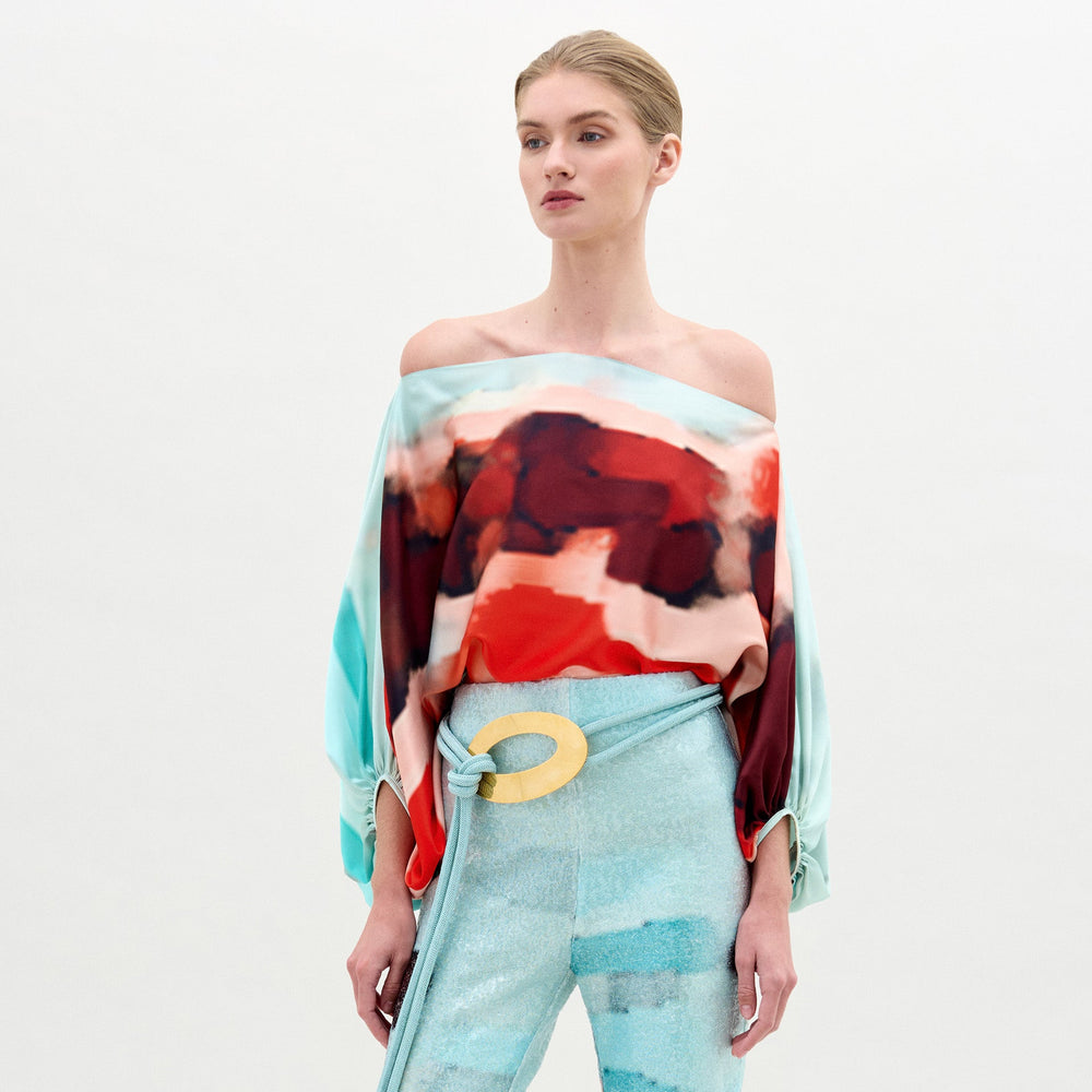 A woman wears the Bellagio Blouse, an off-shoulder red and aqua abstract print silk top with an oversized fit, paired with light blue pants featuring a large circular yellow belt buckle, against a plain background.