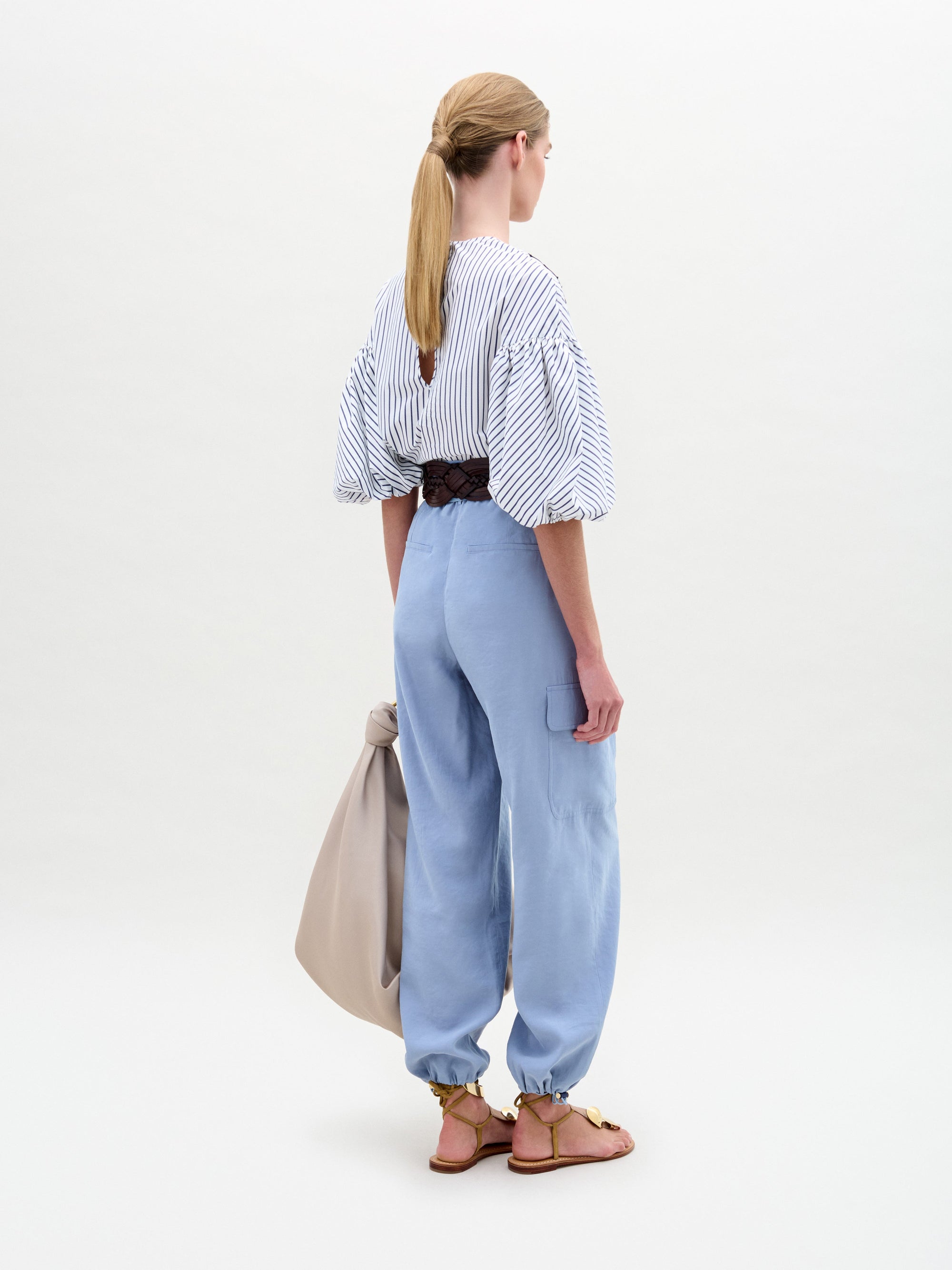 A woman stands against a plain background wearing a striped blouse with blue floral appliqué, the Darnell Pant in light blue, a brown belt with a large buckle, gold sandals, and holding a beige bag.