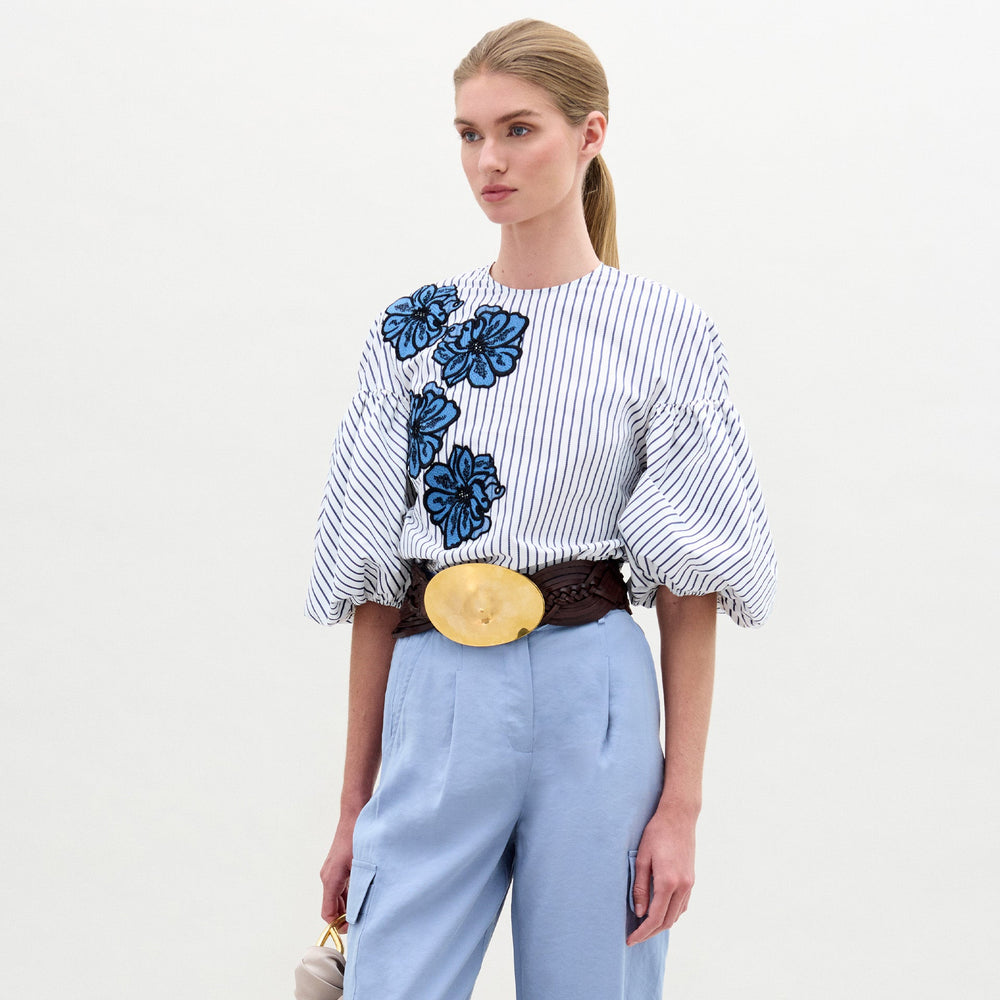 A woman wearing the Belle Blouse, featuring blue floral embroidery and voluminous sleeves, paired with light blue high-waisted pants, a wide brown belt with a large circular buckle, and holding a small handbag.