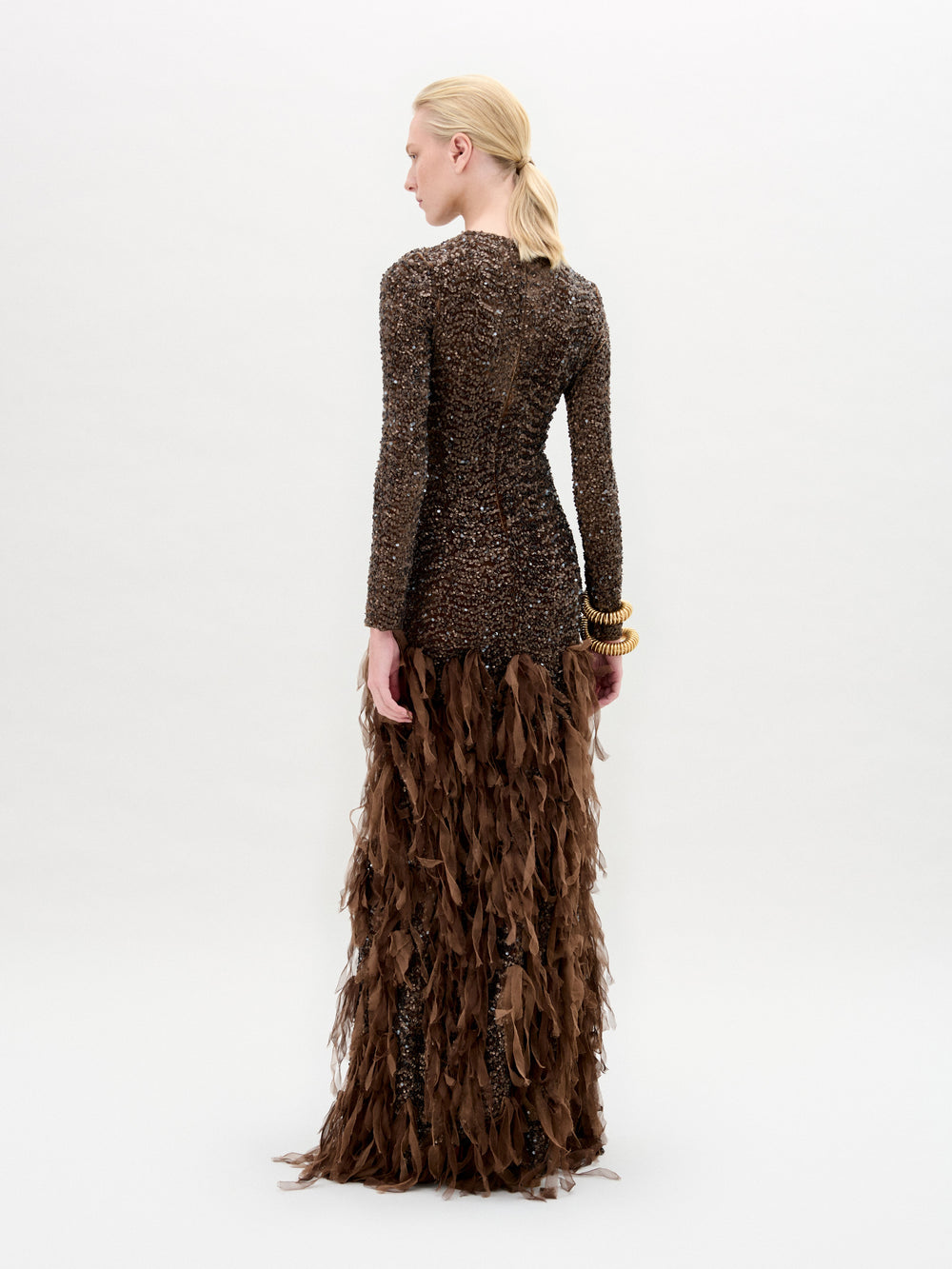 A person stands gracefully in the Bilal Dress, a brown textured maxi with feather-like details made from exquisite Italian fabric, against a plain background.