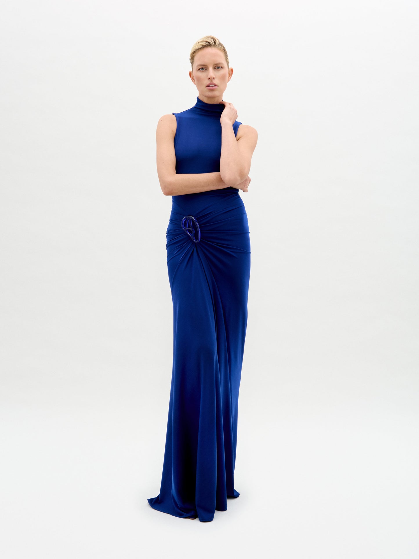 A model wears the Bogna Dress, a sleeveless high-neck blue gown with draped detail, against a plain white background.