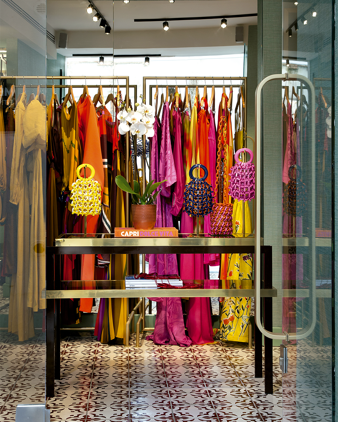 A boutique clothing store displays colorful dresses and handbags on gold racks, with a potted orchid on a central table.