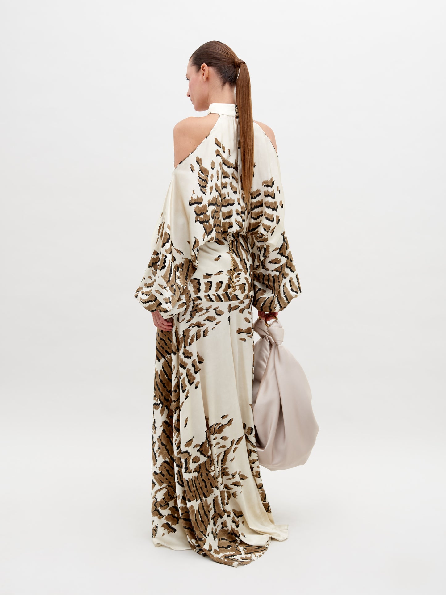 A woman poses in a beige and brown patterned, off-shoulder Bahar Dress Beige Abstract Animal Print holding a large, light beige handbag against a plain white background. This stunning ensemble, available for pre-order now, promises an elegant addition to your wardrobe with an estimated ship date in 2024.