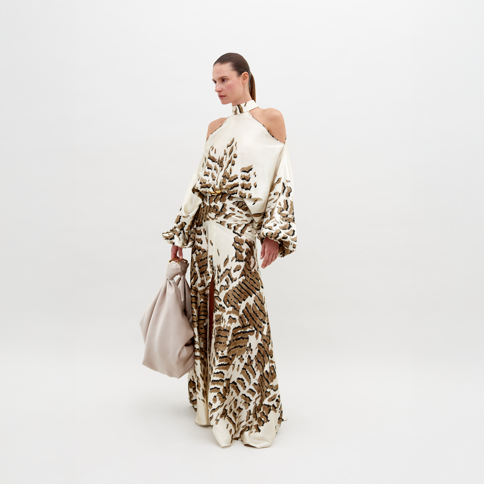 A woman poses in a beige and brown patterned, off-shoulder Bahar Dress Beige Abstract Animal Print holding a large, light beige handbag against a plain white background. This stunning ensemble, available for pre-order now, promises an elegant addition to your wardrobe with an estimated ship date in 2024.