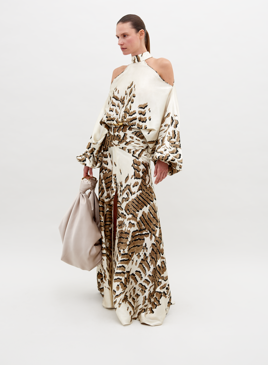 A woman poses in a beige and brown patterned, off-shoulder Bahar Dress Beige Abstract Animal Print holding a large, light beige handbag against a plain white background. This stunning ensemble, available for pre-order now, promises an elegant addition to your wardrobe with an estimated ship date in 2024.