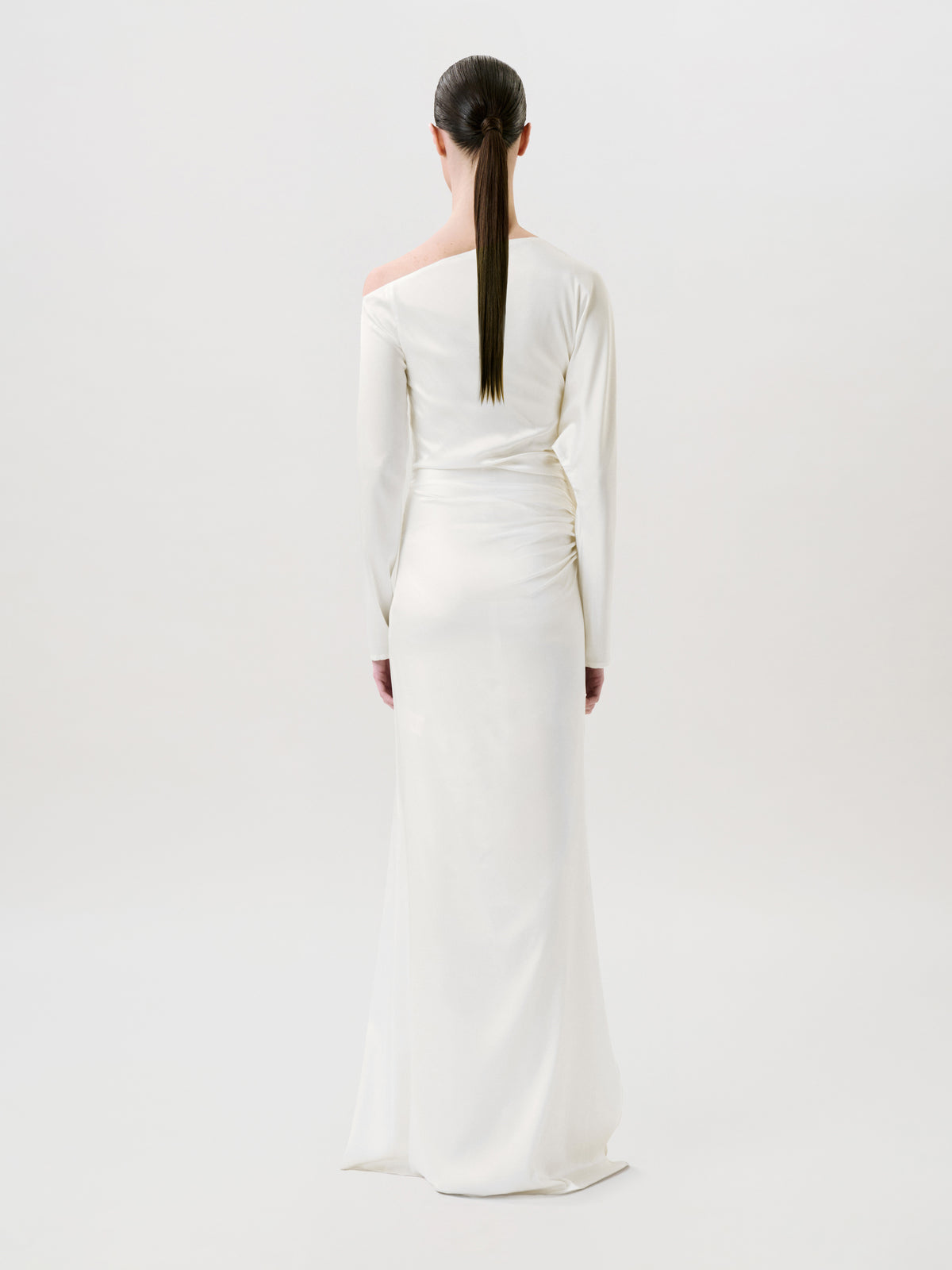A person with long dark hair in a low ponytail wears the floor-length, long-sleeved Bahir Dress in white silk and stands facing away against a plain light background, showcasing timeless elegance in this ready-to-wear piece made in the USA.