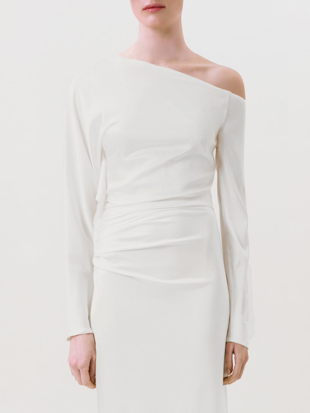 A person models the Bahir Dress—a minimalist white silk, one-shoulder long-sleeve design—standing against a plain background. The ready-to-wear piece is displayed in an image cropped from mid-face to mid-thigh.