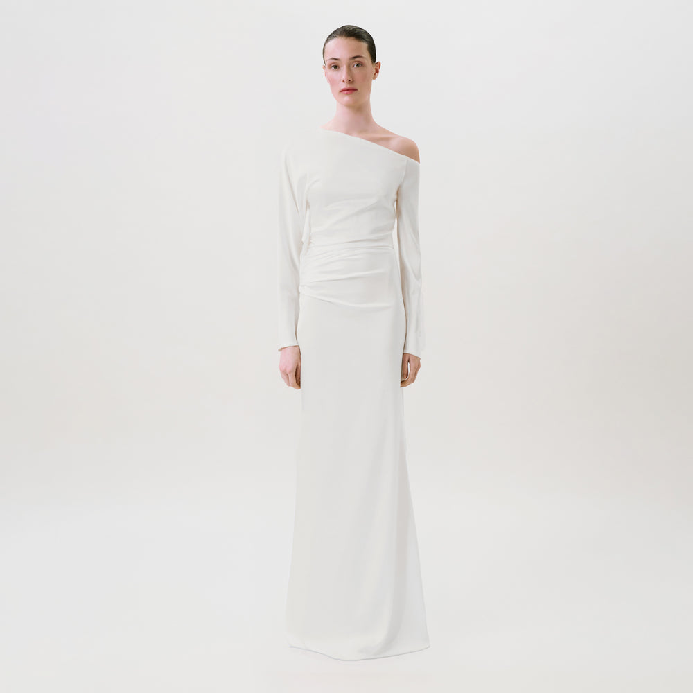 A woman stands against a plain white background wearing the Bahir Dress, a long, white, one-shoulder gown crafted from luxurious silk. This elegant ready-to-wear piece features long sleeves and a draped waist detail, expertly made in the USA for effortless sophistication.