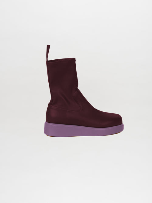 The Baia Boots Lillac are designed with a dark burgundy shade and flat purple sole, made from soft stretch calf leather, presenting a fashion-forward appeal against a plain white background.