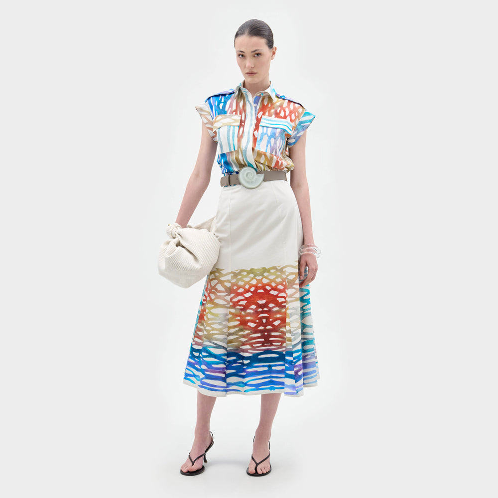 A woman stands against a white background wearing the Erin Skirt, a colorful patterned A-line midi skirt with abstract designs, styled with a matching blouse, wide belt, black sandals, and holding a white bag.