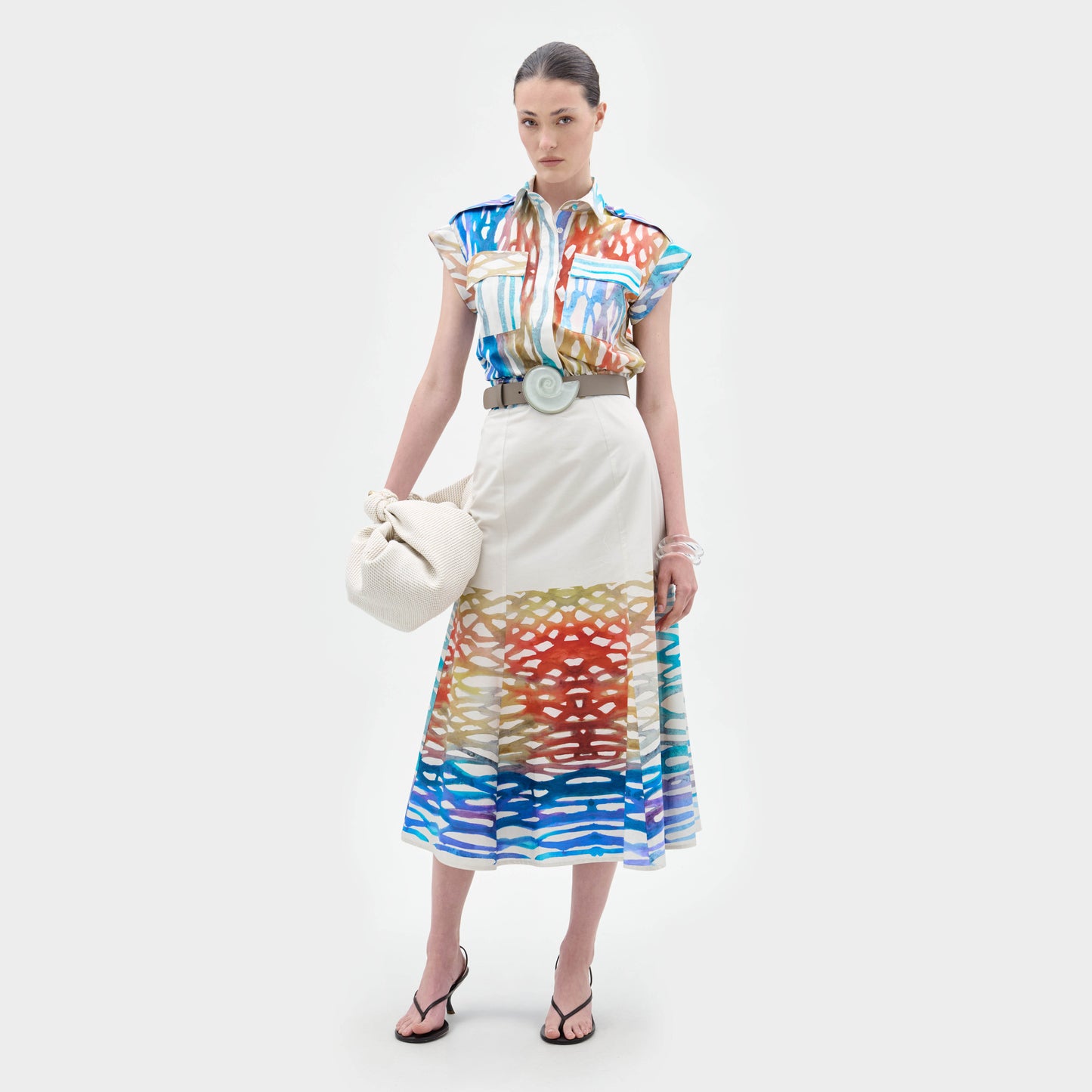 A woman stands against a white background wearing the Erin Skirt, a colorful patterned A-line midi skirt with abstract designs, styled with a matching blouse, wide belt, black sandals, and holding a white bag.