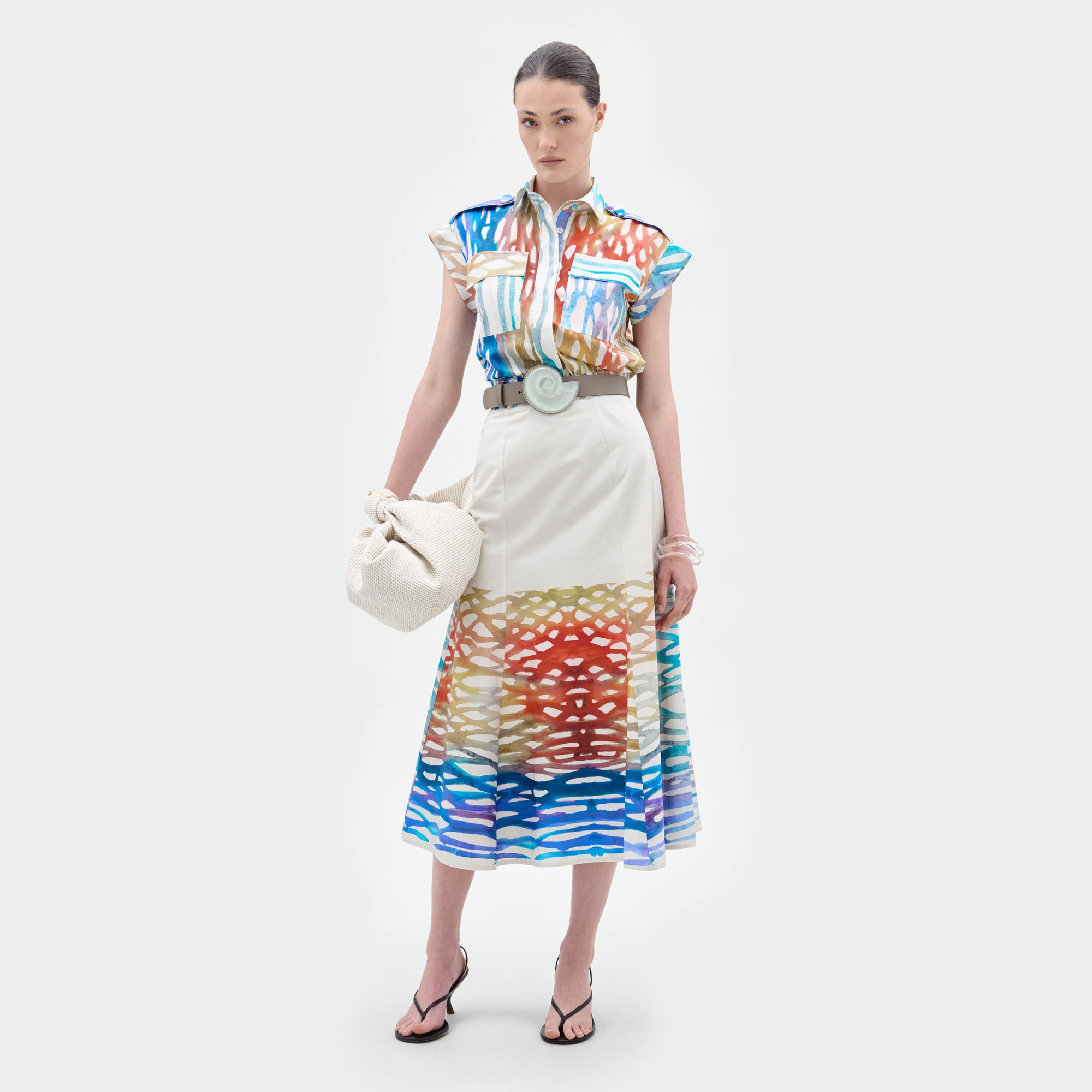 A woman stands against a white background wearing the Erin Skirt, a colorful patterned A-line midi skirt with abstract designs, styled with a matching blouse, wide belt, black sandals, and holding a white bag.