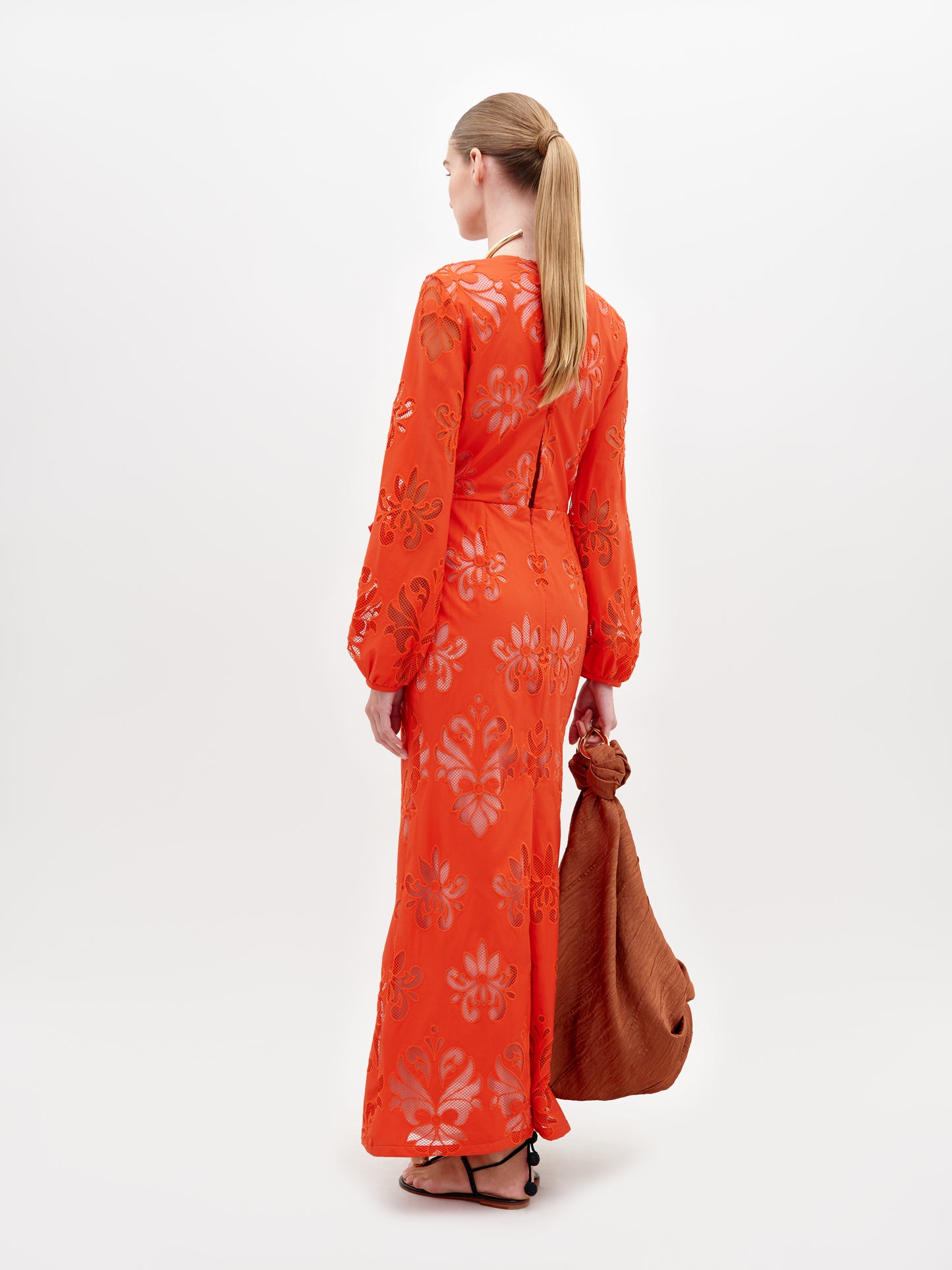 A person stands in a long, bright orange Battia Dress Rouge Embroidery with floral patterns and a front bow tie. They carry a large, rust-colored bag and wear gold jewelry and black sandals. To ensure you receive this stunning ensemble by the desired ship-by date, consider placing your pre-order for 2024 today.