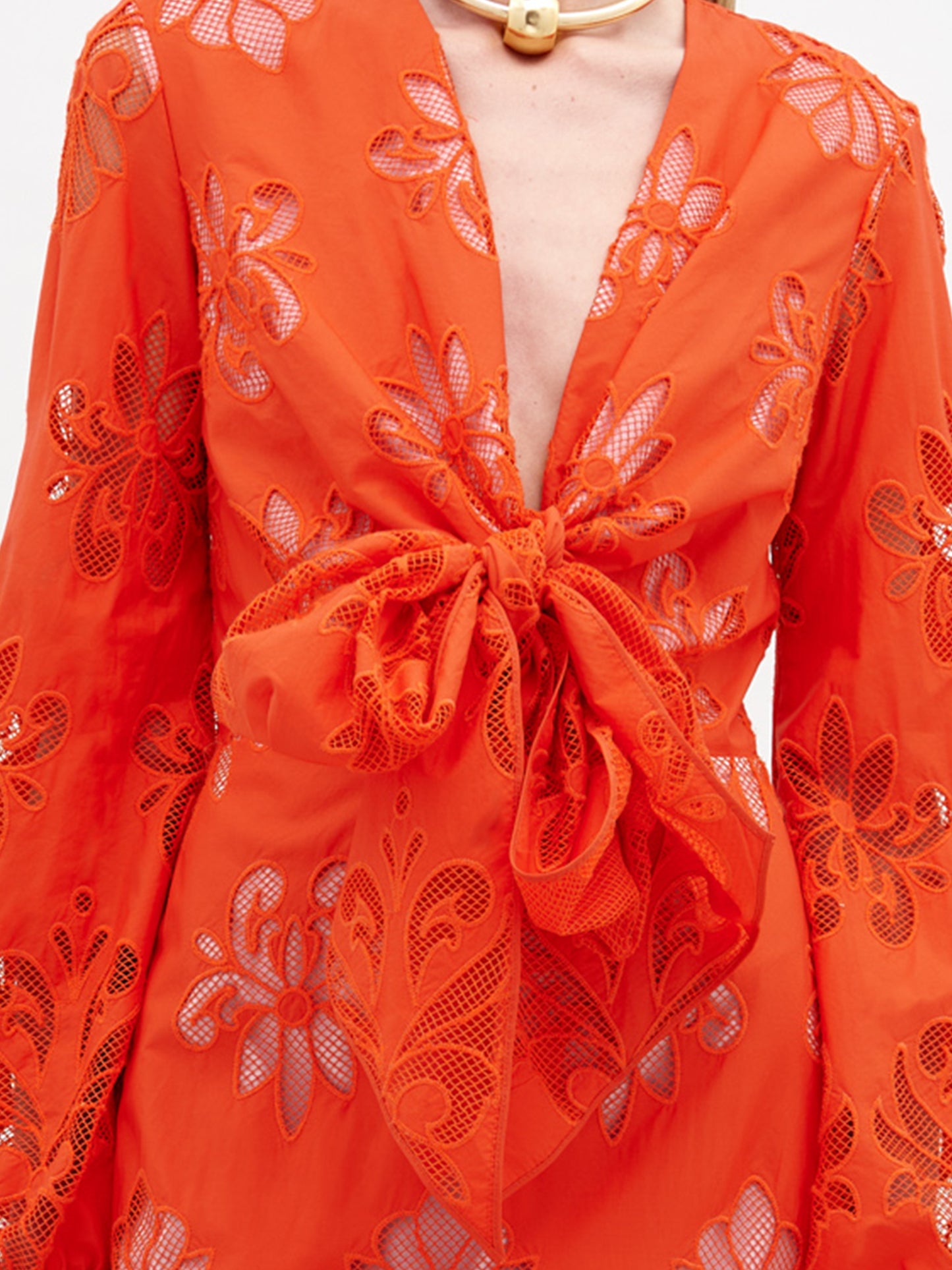 A person stands in a long, bright orange Battia Dress Rouge Embroidery with floral patterns and a front bow tie. They carry a large, rust-colored bag and wear gold jewelry and black sandals. To ensure you receive this stunning ensemble by the desired ship-by date, consider placing your pre-order for 2024 today.