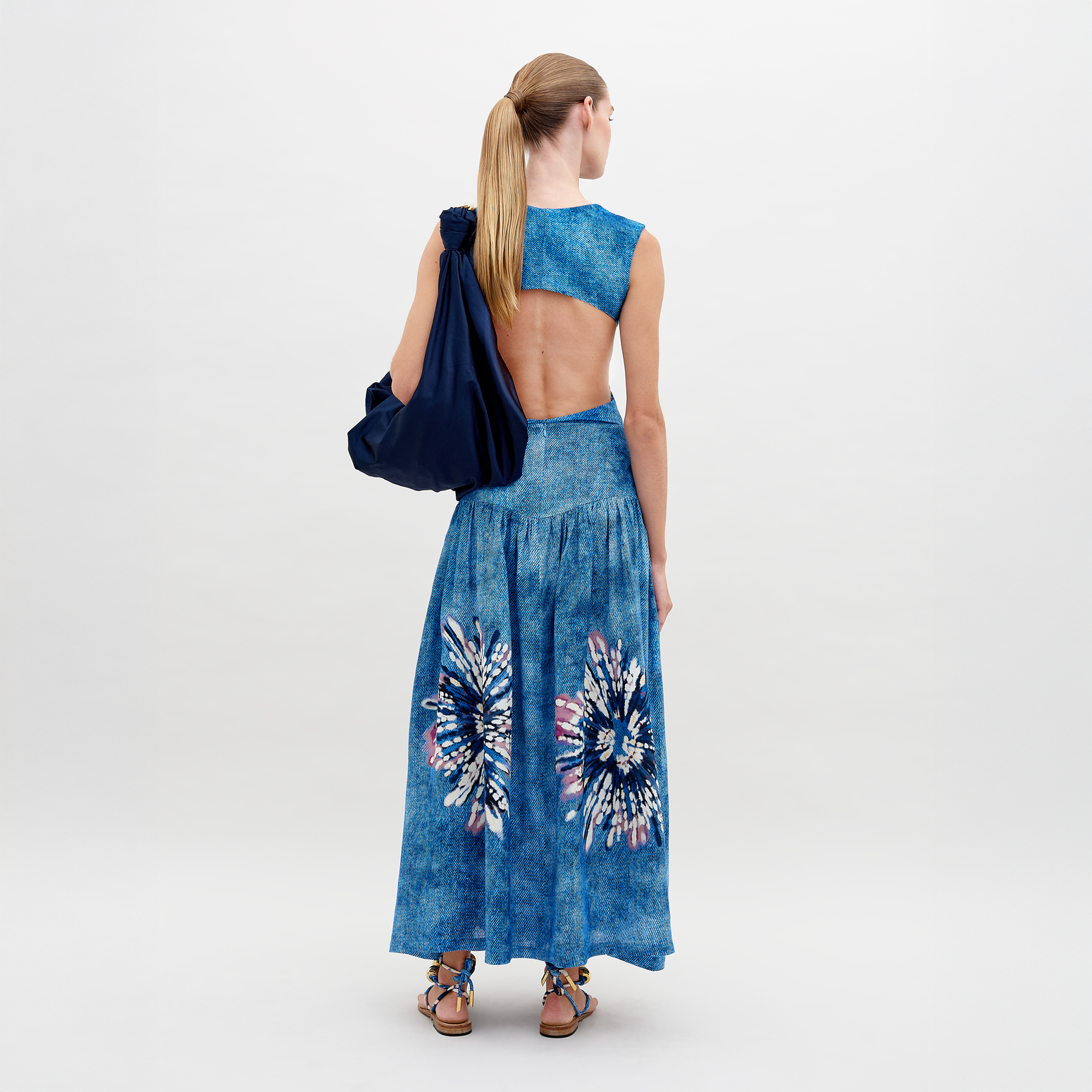 A woman confidently poses against a white background in a blue backless dress with floral patterns, paired with the Abla Skirt featuring an abstract print, and holds a navy bag over her shoulder.