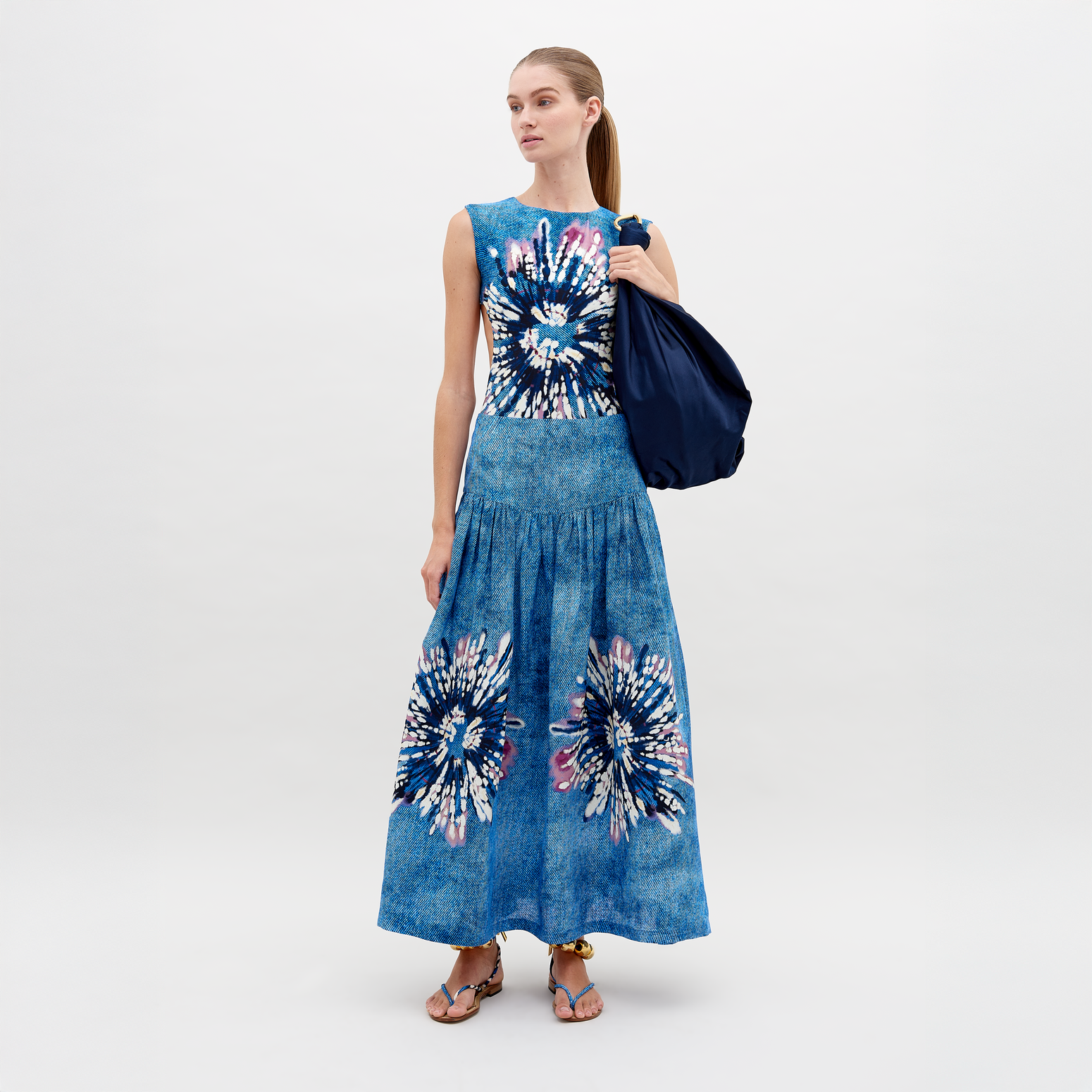 A woman confidently poses against a white background in a blue backless dress with floral patterns, paired with the Abla Skirt featuring an abstract print, and holds a navy bag over her shoulder.