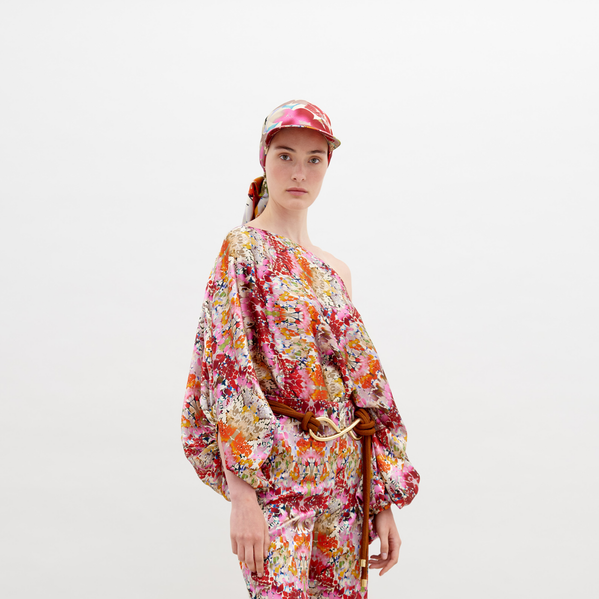A person stands, wearing a colorful floral outfit with a matching cap. The Bellagio Blouse Multicolor Animal Print has one shoulder exposed, long billowing sleeves, and is paired with matching pants and a knotted belt. Pre-order now to ensure it ships by November 15th, 2024.
