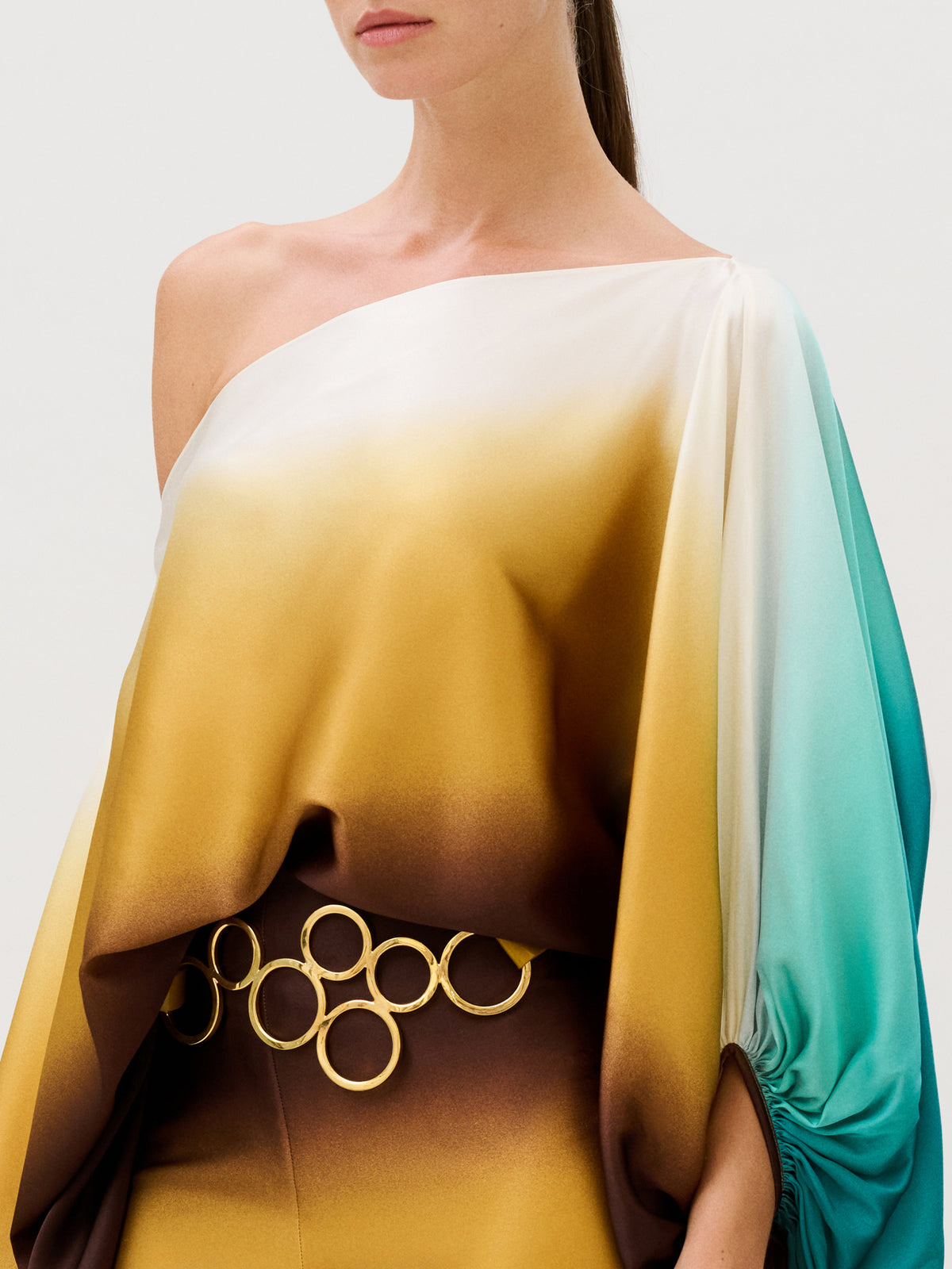 Bellagio Blouse: a one-shoulder Italian silk blouse featuring a gradient print of white, mustard, and teal, worn with a belt made of interlocking gold-tone rings.