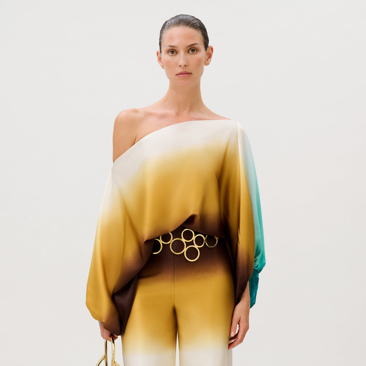 Model wearing the Bellagio Blouse, an off-the-shoulder, ombre-toned Italian silk top with one long, loose sleeve and a gold-tone circular link belt at the waist.