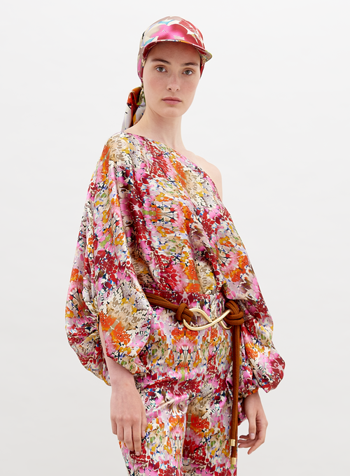 A person stands, wearing a colorful floral outfit with a matching cap. The Bellagio Blouse Multicolor Animal Print has one shoulder exposed, long billowing sleeves, and is paired with matching pants and a knotted belt. Pre-order now to ensure it ships by November 15th, 2024.