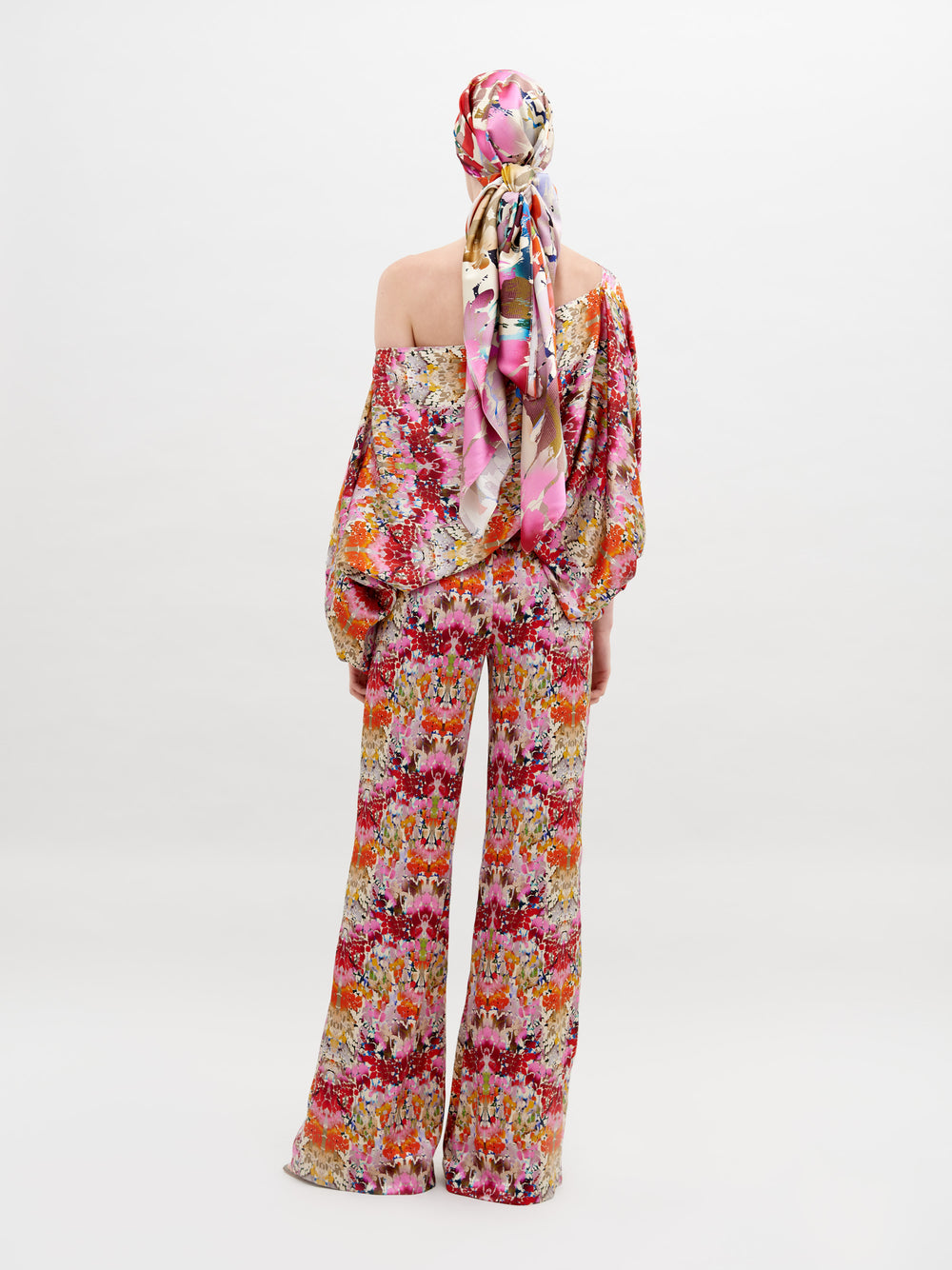 Person wearing a floral-patterned outfit consisting of an Andie Pant Multicolor Animal Print, a one-shoulder top, and a matching cap, standing against a plain white background. This stunning ensemble is now available for pre-order with an expected ship date of November 15th, 2024.