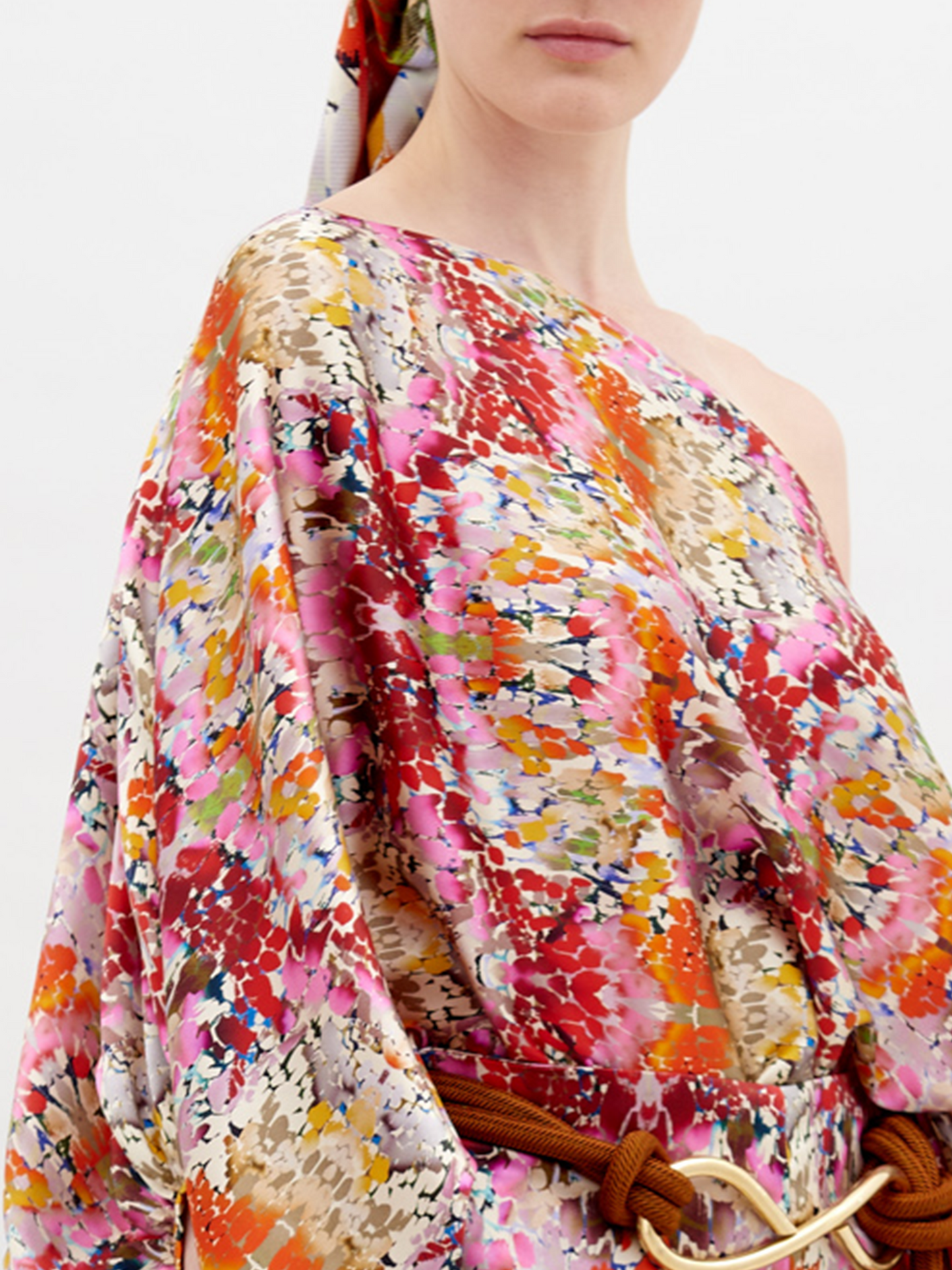 A person stands, wearing a colorful floral outfit with a matching cap. The Bellagio Blouse Multicolor Animal Print has one shoulder exposed, long billowing sleeves, and is paired with matching pants and a knotted belt. Pre-order now to ensure it ships by November 15th, 2024.