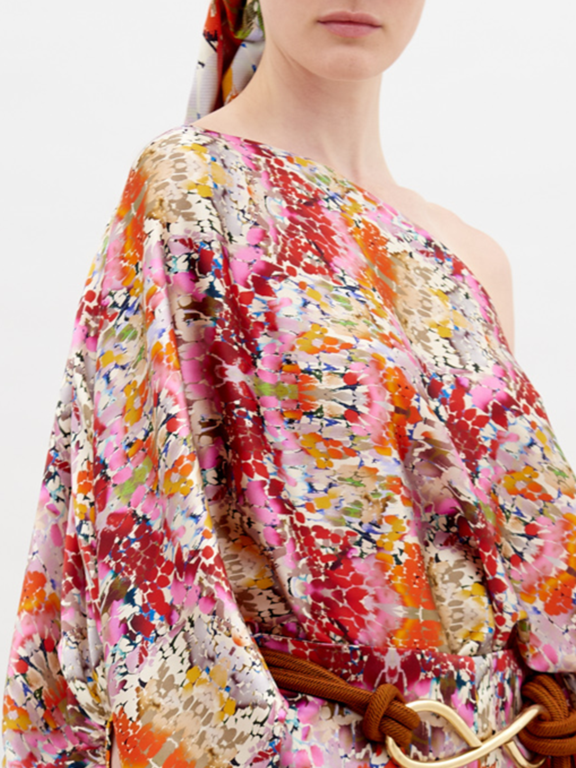 A person stands, wearing a colorful floral outfit with a matching cap. The Bellagio Blouse Multicolor Animal Print has one shoulder exposed, long billowing sleeves, and is paired with matching pants and a knotted belt. Pre-order now to ensure it ships by November 15th, 2024.