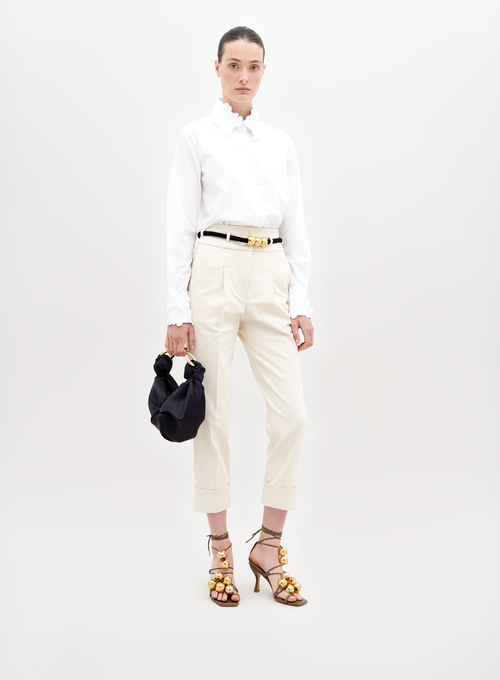 A person stands against a plain background, wearing a white blouse, Beryl Pant Beige, black belt, and golden embellished heels, holding a black handbag with a knot detail. This stylish ensemble is perfect for those who pre-order now to ensure it arrives by the November 15th, 2024 ship-by date.