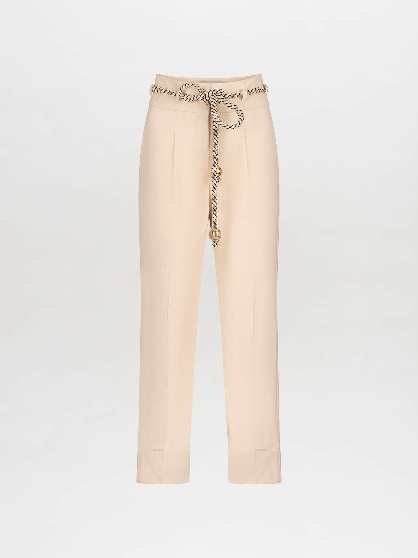 A person stands against a plain background, wearing a white blouse, Beryl Pant Beige, black belt, and golden embellished heels, holding a black handbag with a knot detail. This stylish ensemble is perfect for those who pre-order now to ensure it arrives by the November 15th, 2024 ship-by date.