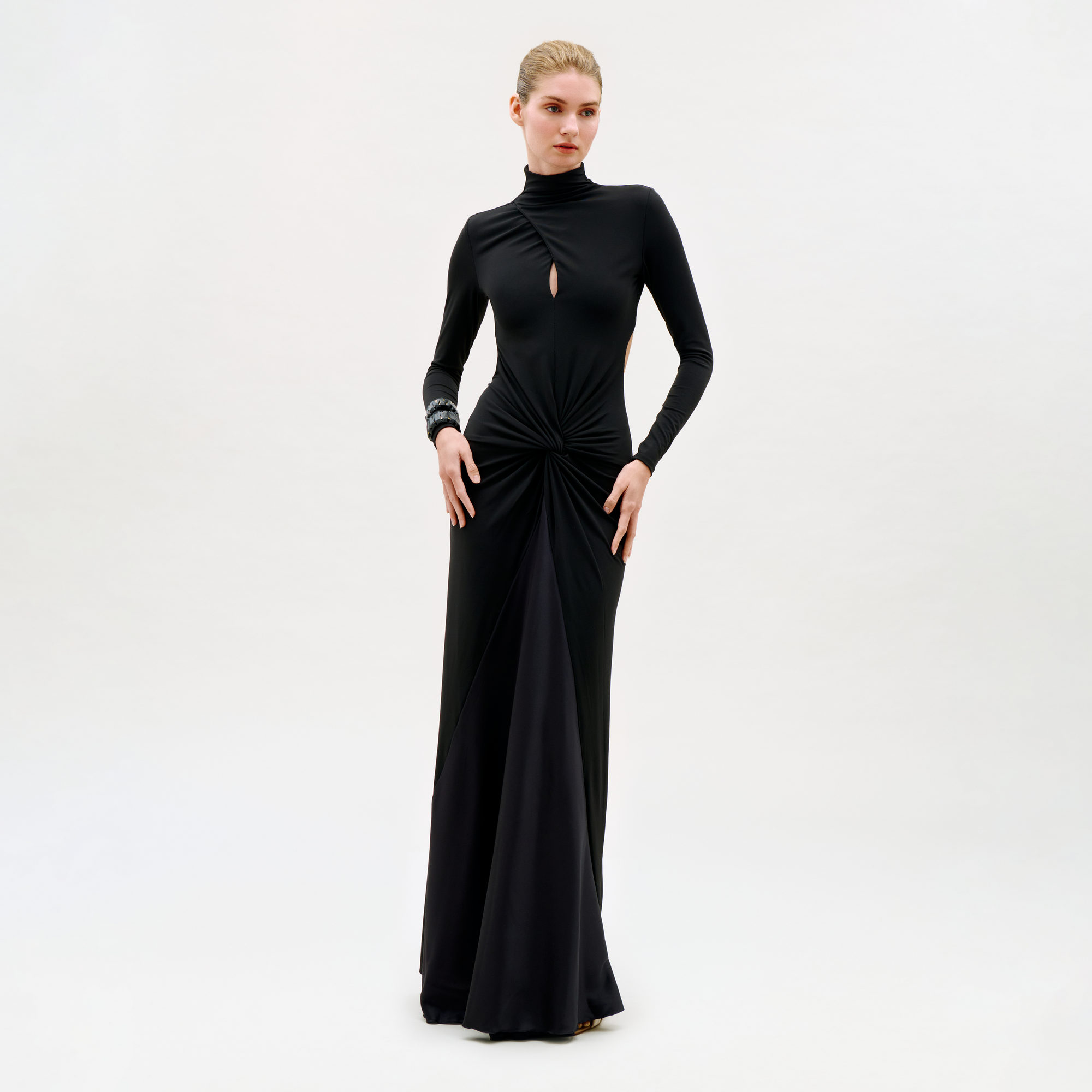 A woman wears the Birdy Dress—a long-sleeved, floor-length black gown featuring a small front keyhole cutout and gathered waist—standing against a plain, light background.