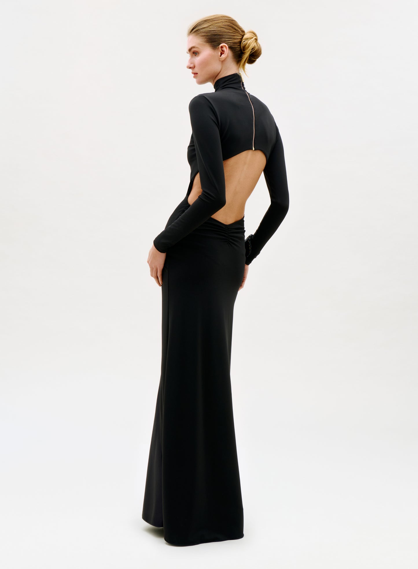A person stands against a plain white background wearing the Birdy Dress, a long, fitted black gown featuring long sleeves and an open back, with their hair styled in a neat bun.
