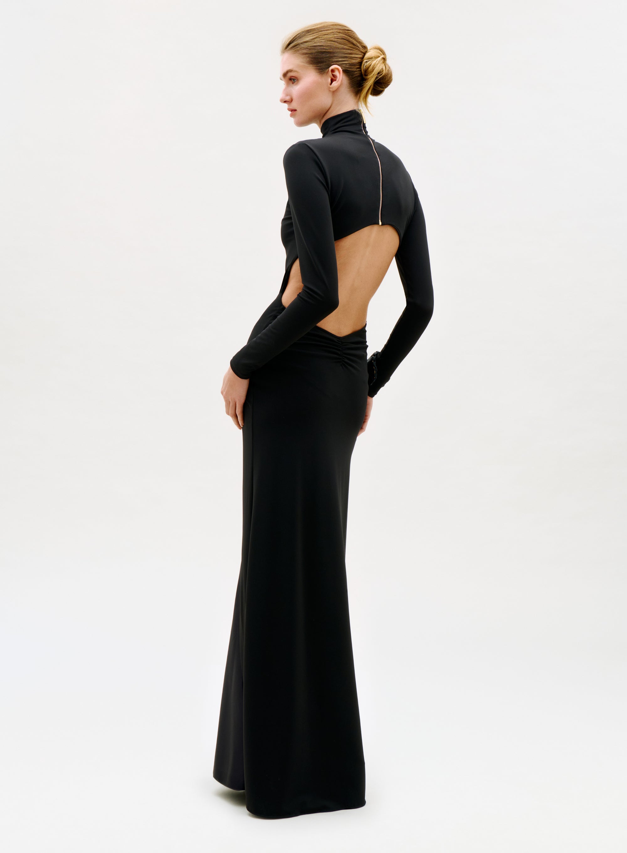 A person stands against a plain white background wearing the Birdy Dress, a long, fitted black gown featuring long sleeves and an open back, with their hair styled in a neat bun.