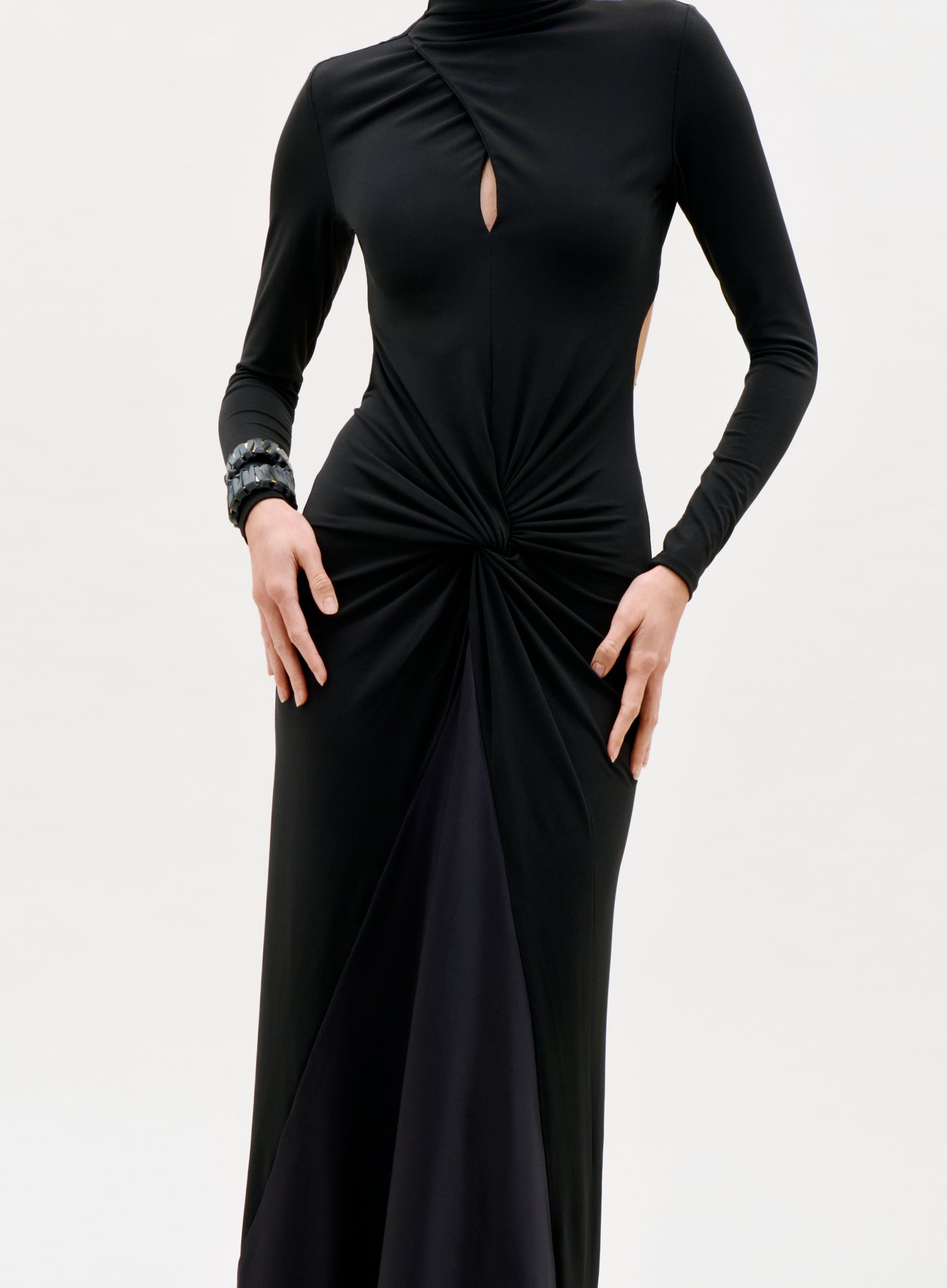A person wearing the Birdy Dress, a long-sleeved black dress featuring a keyhole chest cutout and gathered twist at the waist, with a bracelet on one wrist, stands against a plain light background.
